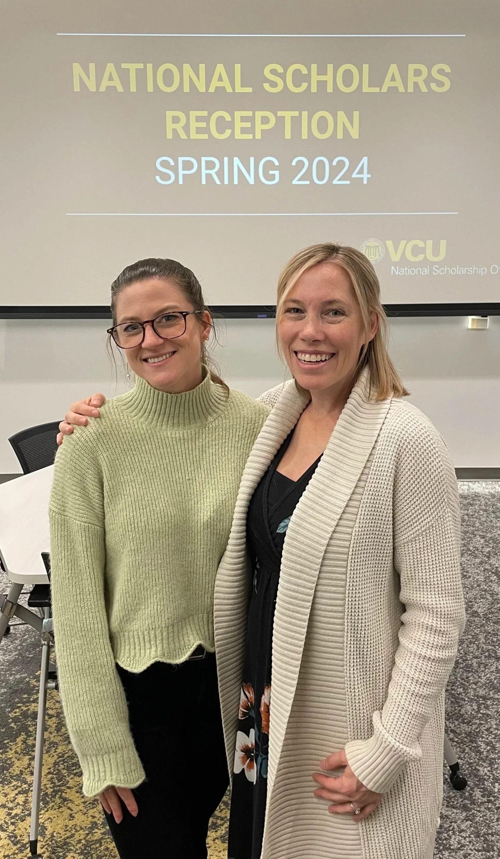 Two women smiling and standing close together at a formal event. Behind them is a projector screen with the text 'National Scholars Reception Spring 2024' and a logo for VCU.