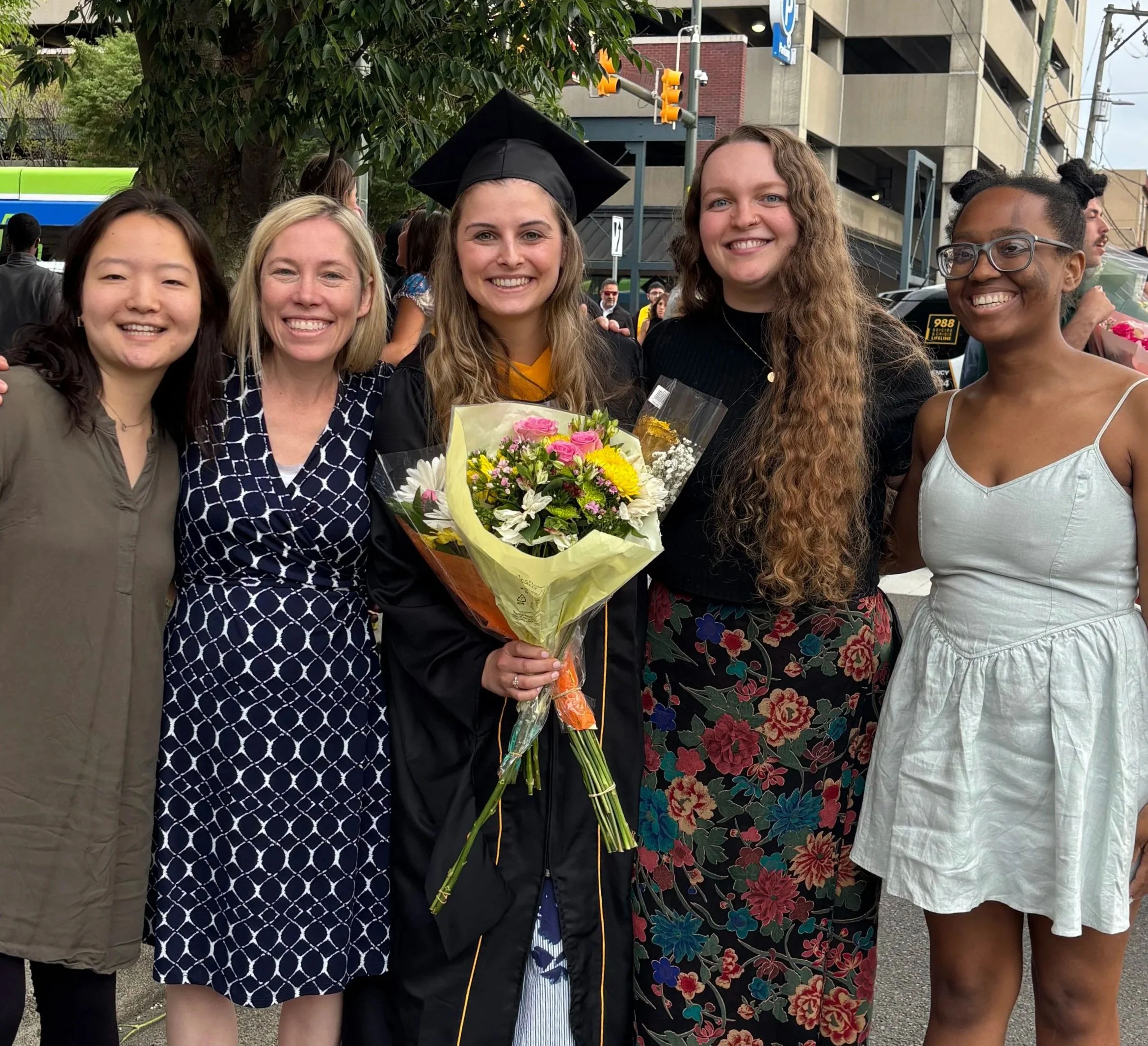 A group of five diverse women standing outdoors, with the center woman wearing a graduation cap and gown holding a bouquet of flowers, smiling at a graduation ceremony.