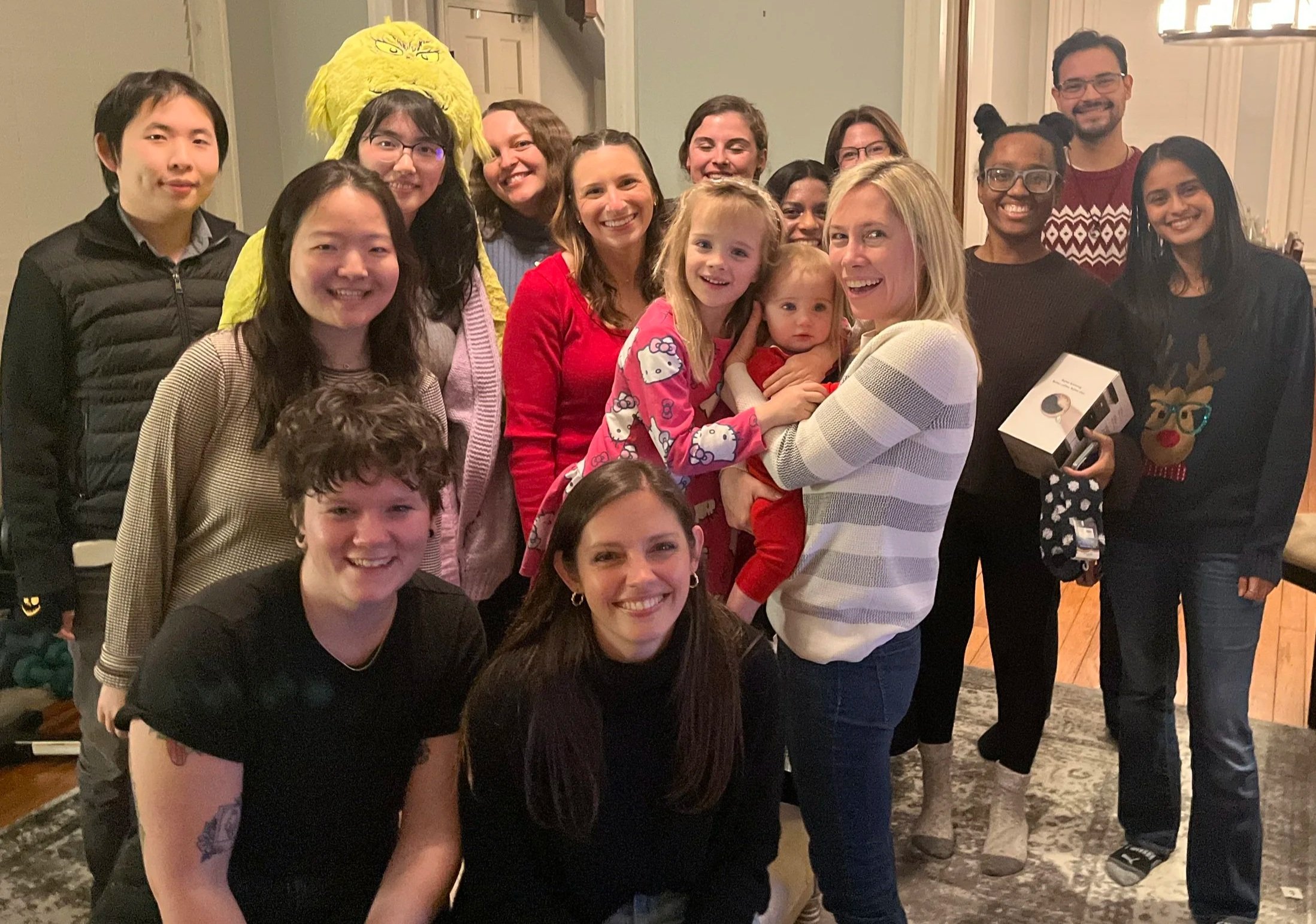 Group of people gathered together, smiling for a photo, some wearing festive accessories like a Grinch hat and reindeer antlers, in a room with wooden floors and decorated for a celebration.