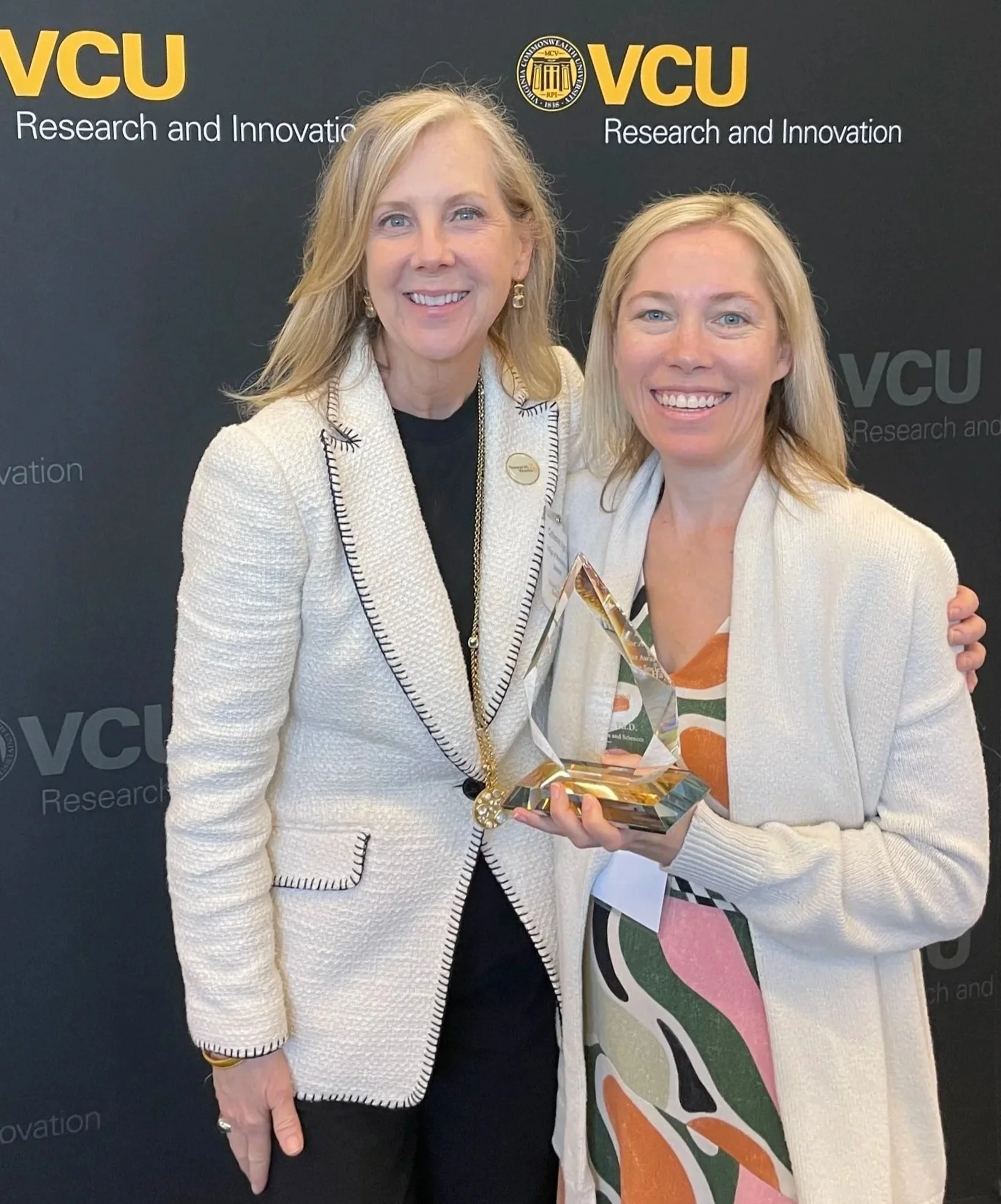 Two women smiling, standing in front of a black backdrop with yellow and white text that reads 'VCU Research and Innovation' and the VCU logo. One woman is holding a glass award.