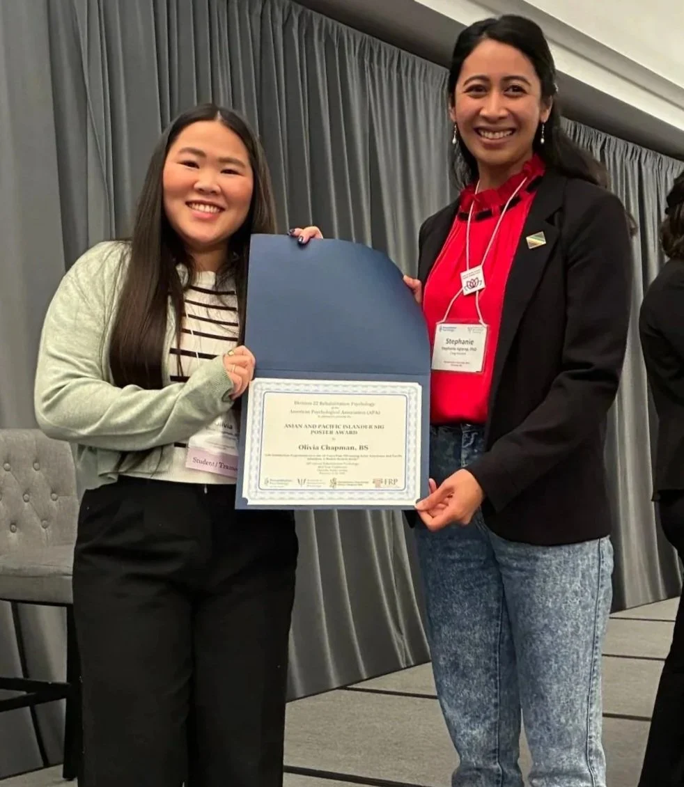 Two women smiling, one holding a certificate inside a blue folder, at an indoor event with gray curtains in the background.