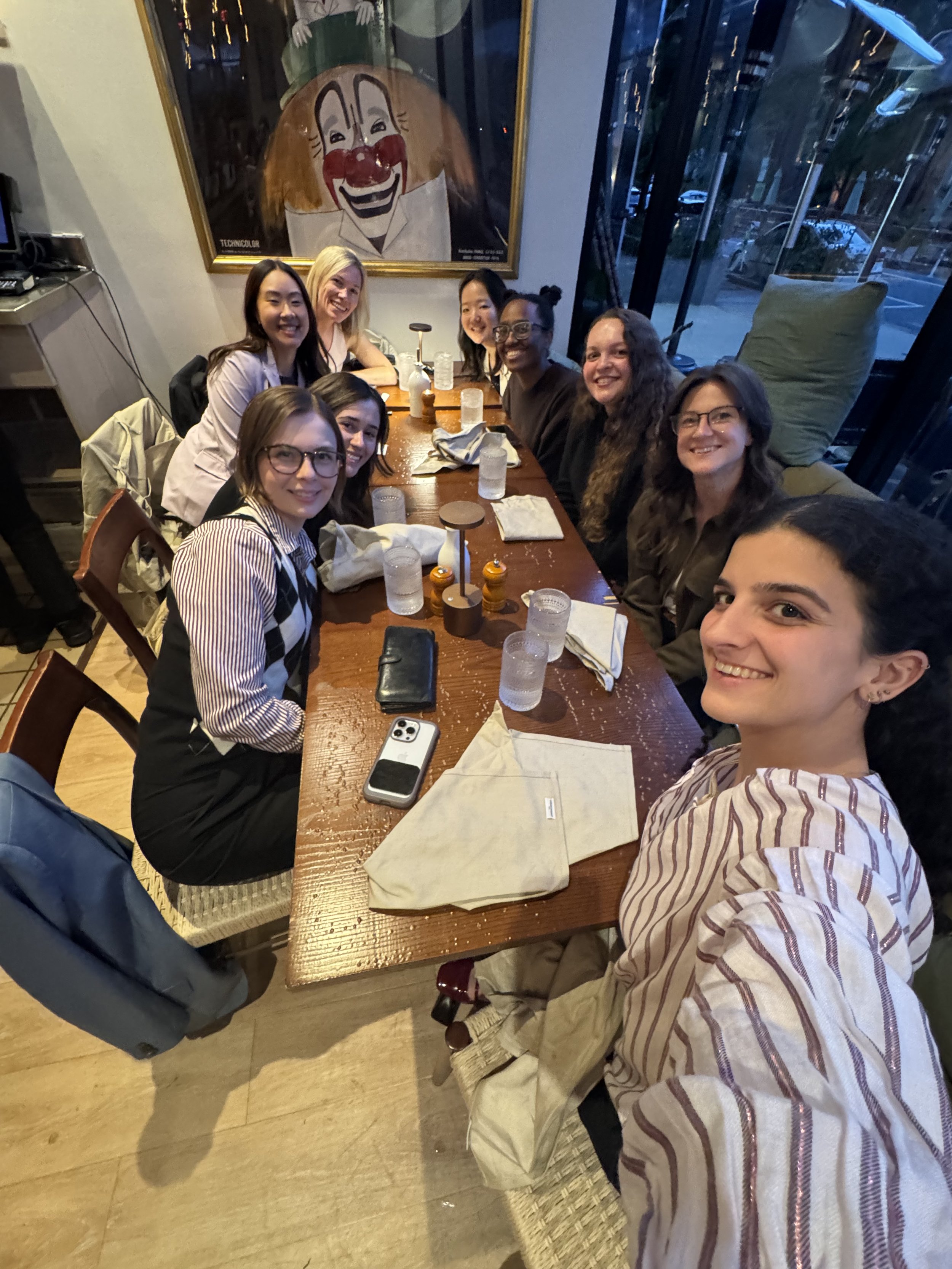 A group of eight diverse women sitting around a wooden dining table in a restaurant, smiling at the camera. The table has glasses of water, napkins, and salt and pepper shakers. Behind them is a large framed clown face painting on the wall, with larg