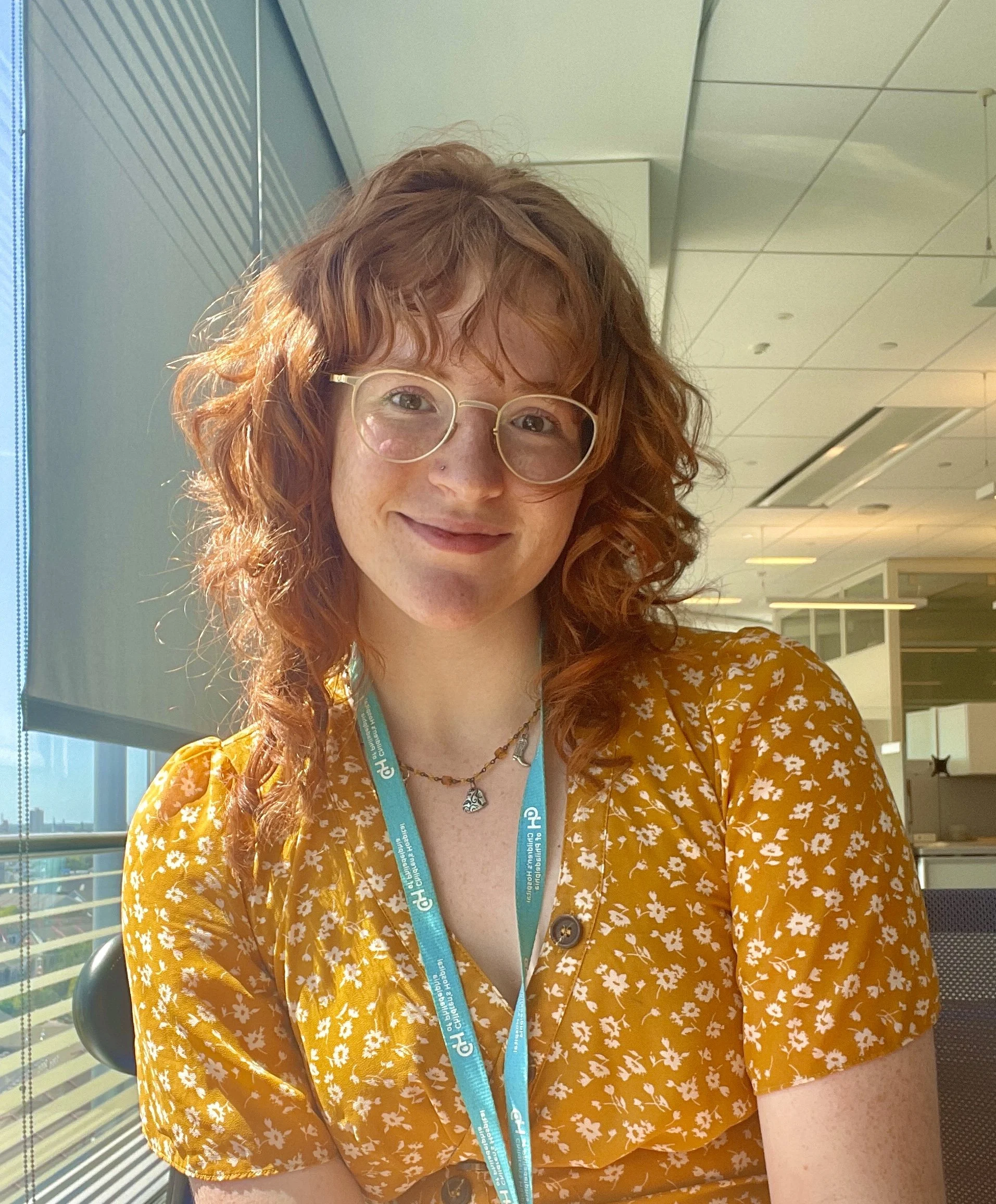 Sammi Weiss. A woman with red curly hair, wearing glasses and a yellow floral blouse, sitting indoors near a large window with blinds, smiling at the camera.