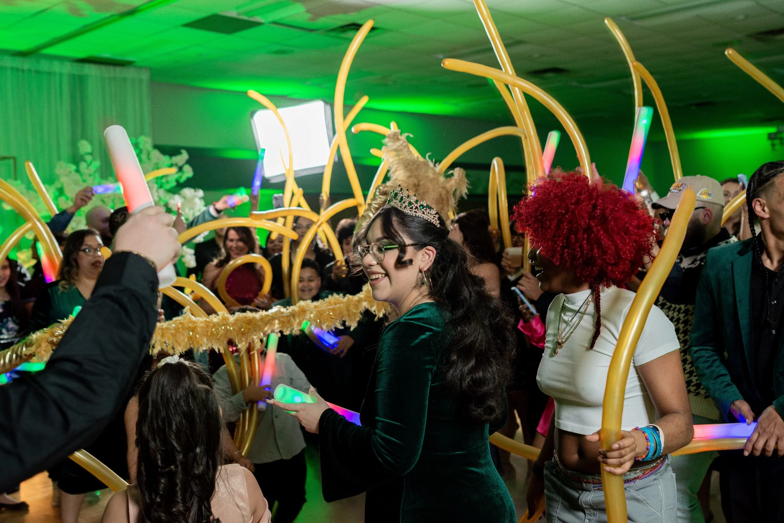 A crowd of people at a celebration or party, with some dancing and holding glow sticks, wearing fun costumes and accessories, in a decorated indoor venue with green lighting.