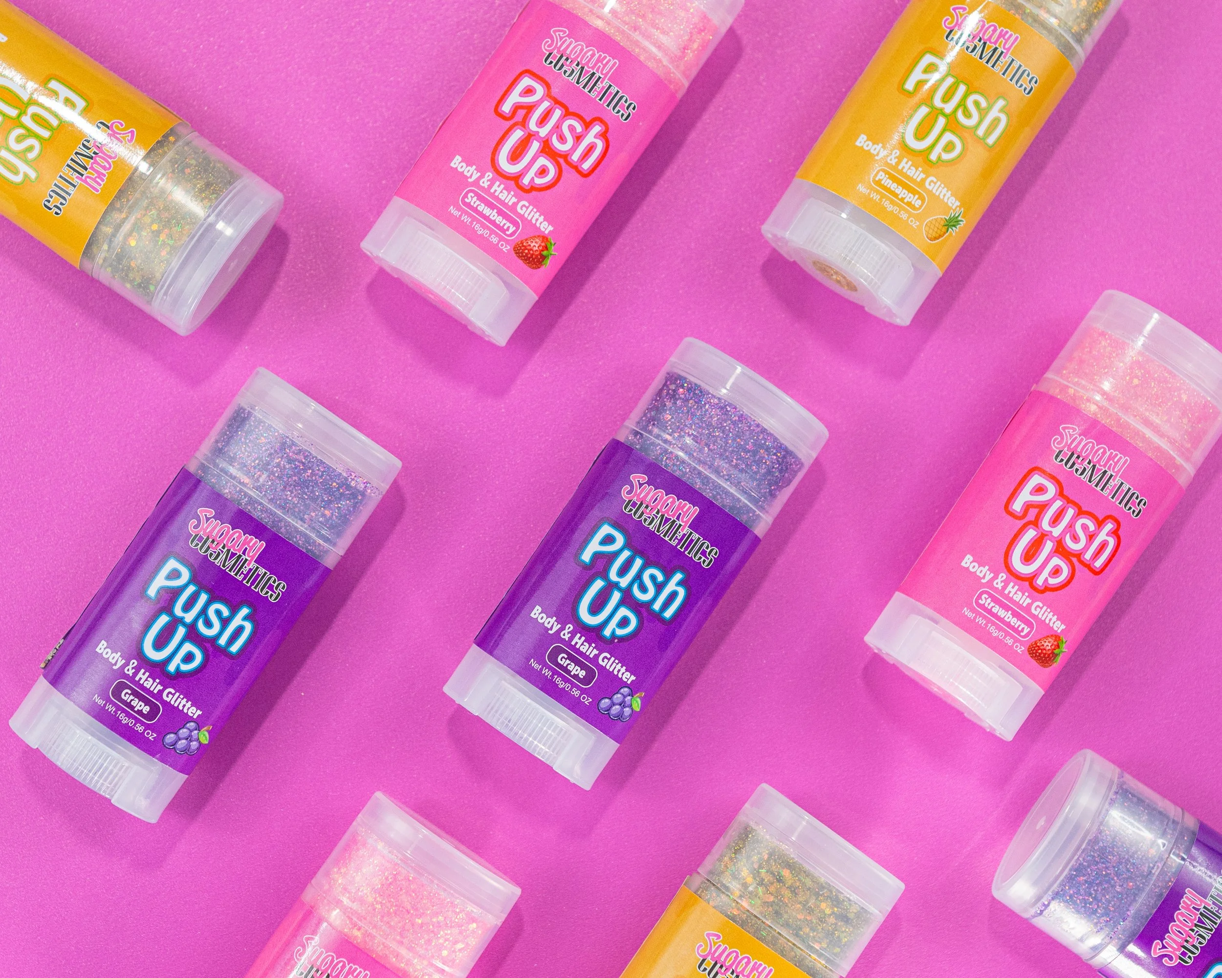 Multiple containers of Sugary Cosmetics Push Up Body & Hair Glitter in various colors including pink, purple, and yellow, arranged on a pink background.