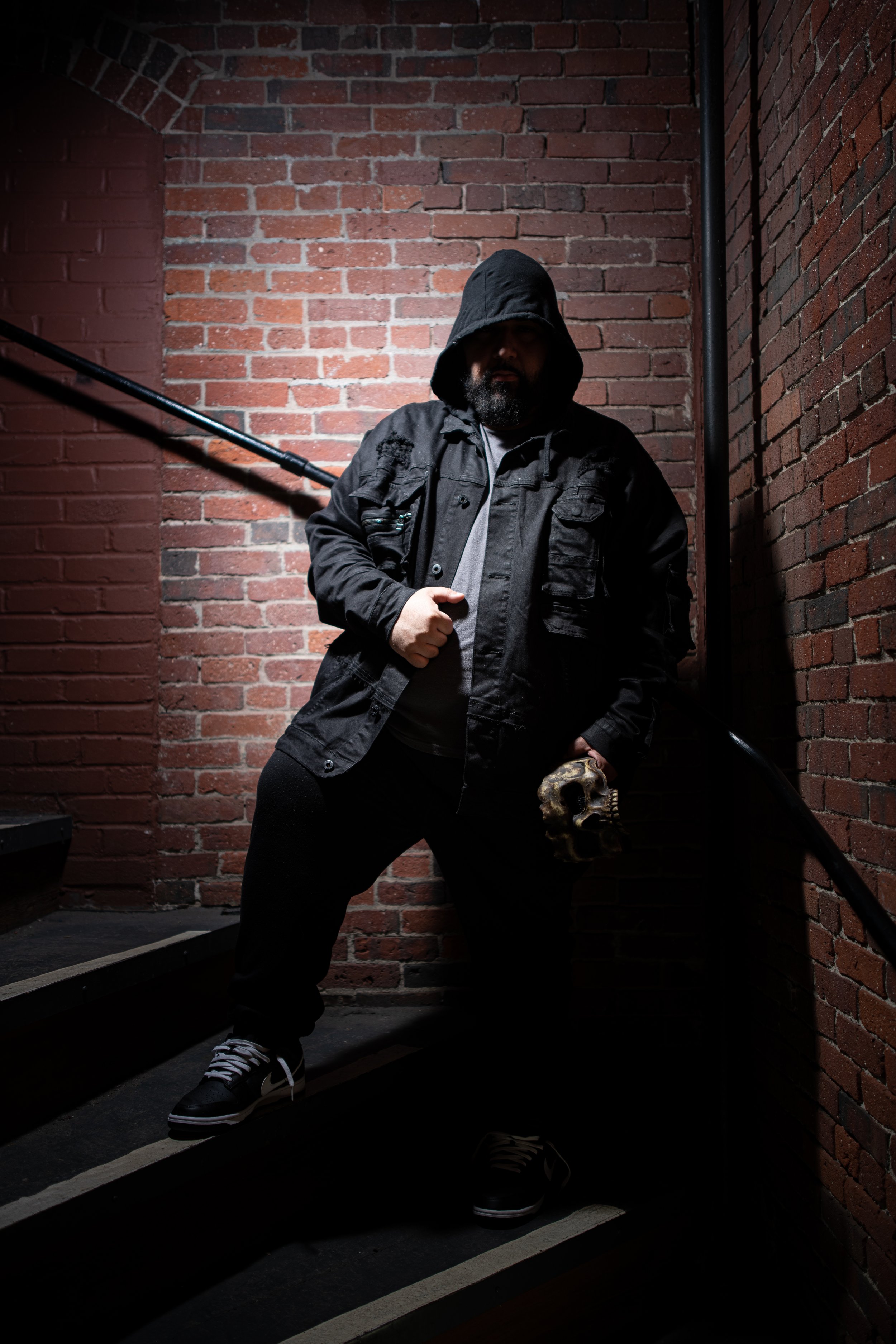 A man wearing a black hoodie and jacket standing on a staircase against a brick wall, holding a mask