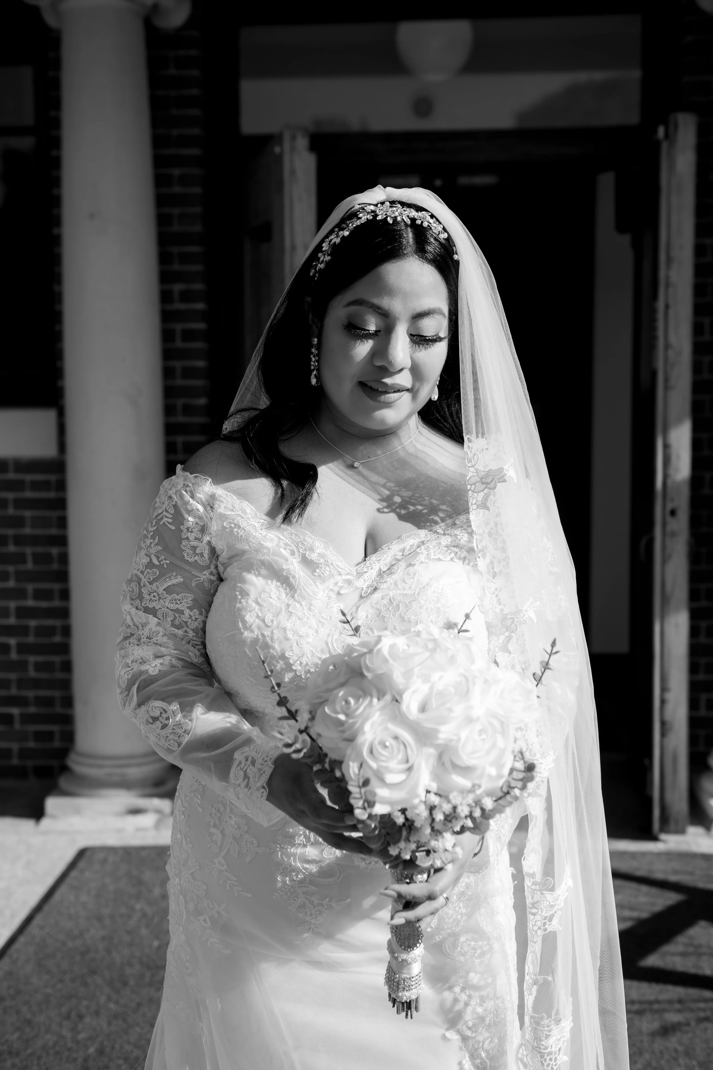 Black and white photo of a bride in a lace wedding dress holding a bouquet of roses outside a building.