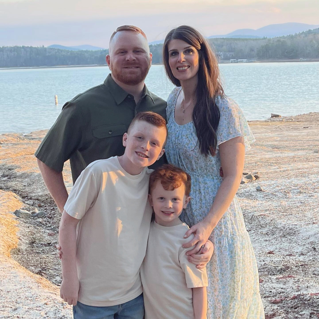 A family of four enjoying a lakeside sunset. The father has short light hair and a beard, wearing a dark green button-up shirt. The mother has long dark hair, wearing a light-colored, floral dress. Their two sons, both with red hair, are in cream-colored shirts, standing in front of them. The background shows a lake, distant trees, and mountains.