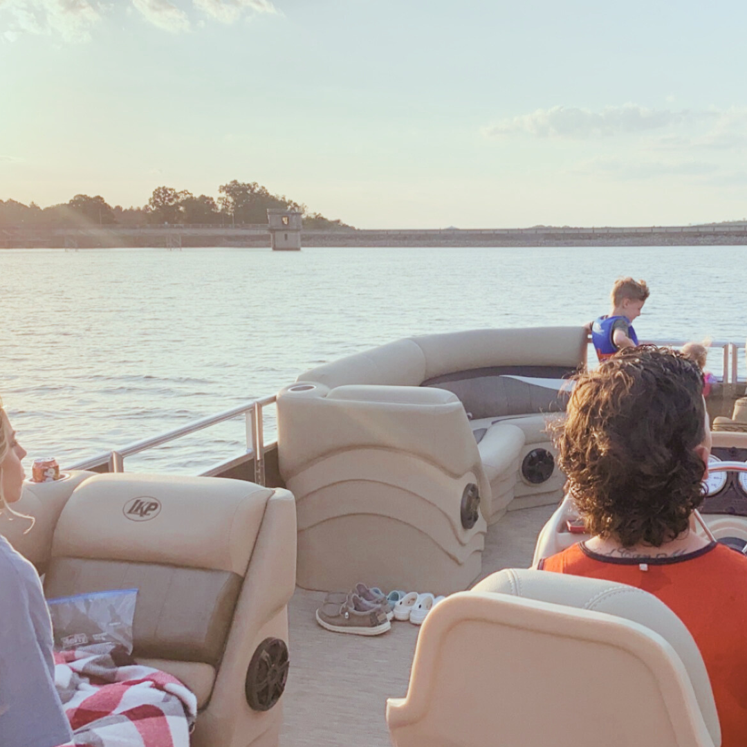 People relaxing on a boat with beige seats, floating on a body of water with a dam and trees in the background, during sunset.