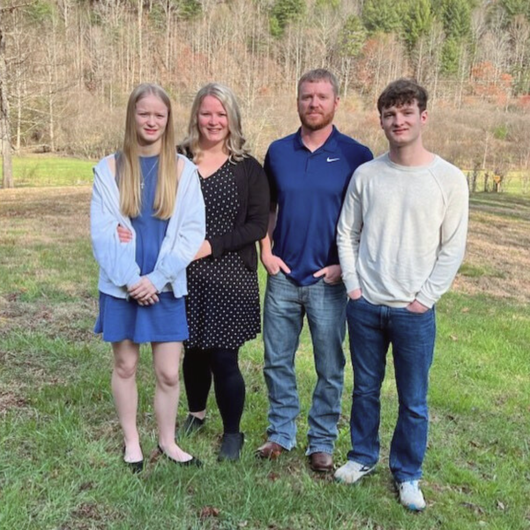 A family of four standing outside on a grassy field with trees in the background, posing for a photo.