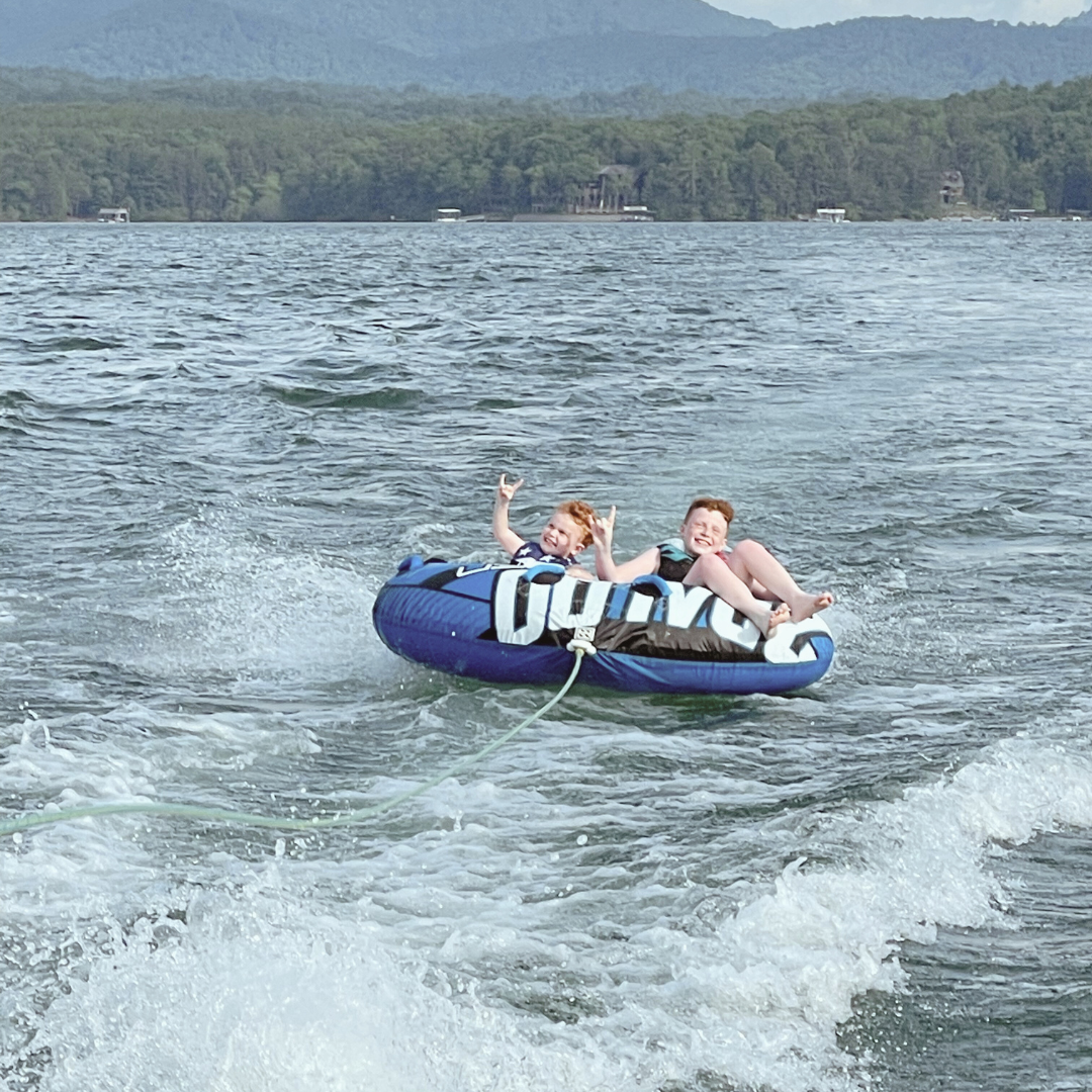 Two children on an inflatable tube being pulled by a boat on a lake, smiling and making peace signs with their hands.