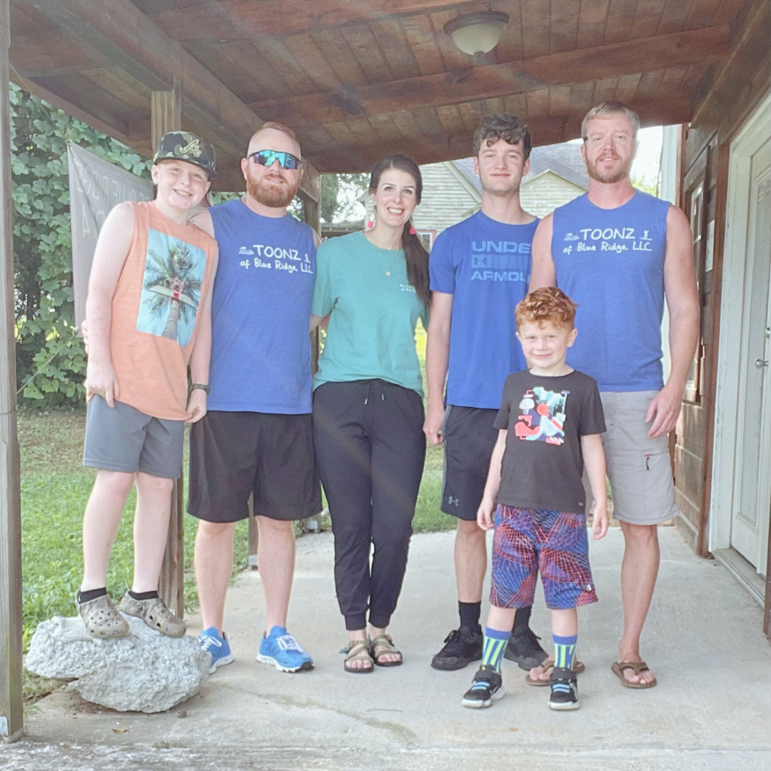 Family group photo outdoors on a porch with six people, including children and adults, smiling and standing close together.