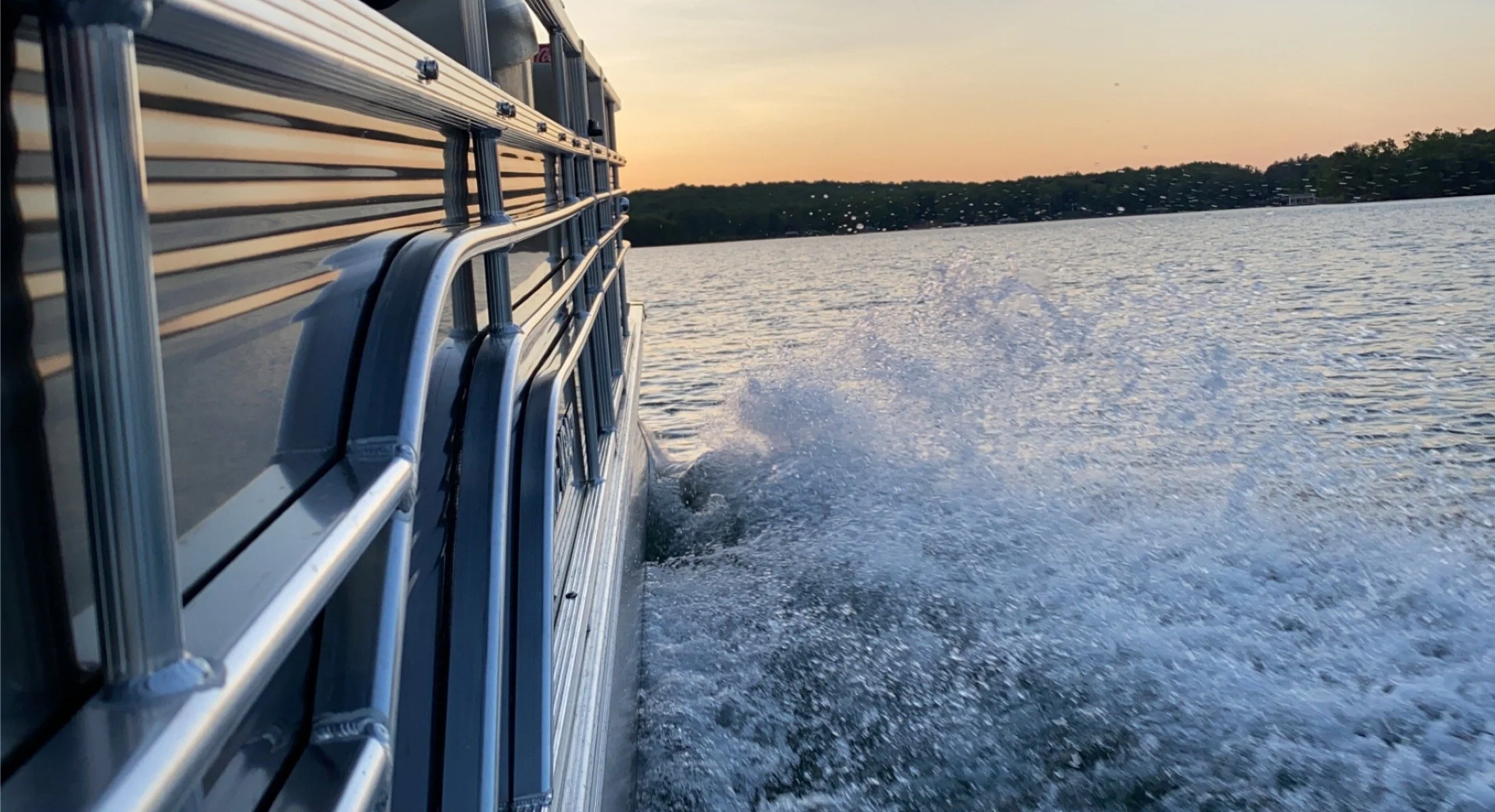 View from a boat moving on a lake at sunset, with water splashing behind and a distant shoreline with trees.