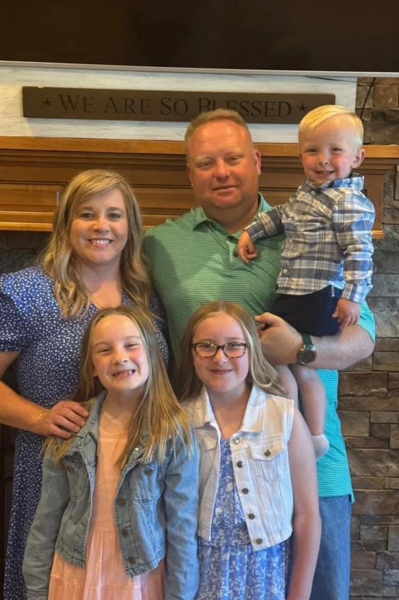 Family of five posing indoors in front of a fireplace with a sign that reads 'We are so blessed' on the mantel, smiling at the camera.