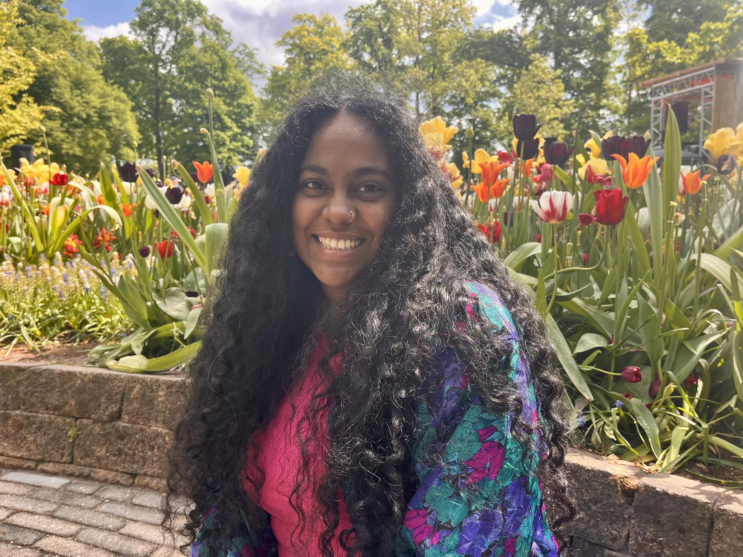 A woman with long curly black hair smiling outdoors in front of a colorful flower bed at a park.