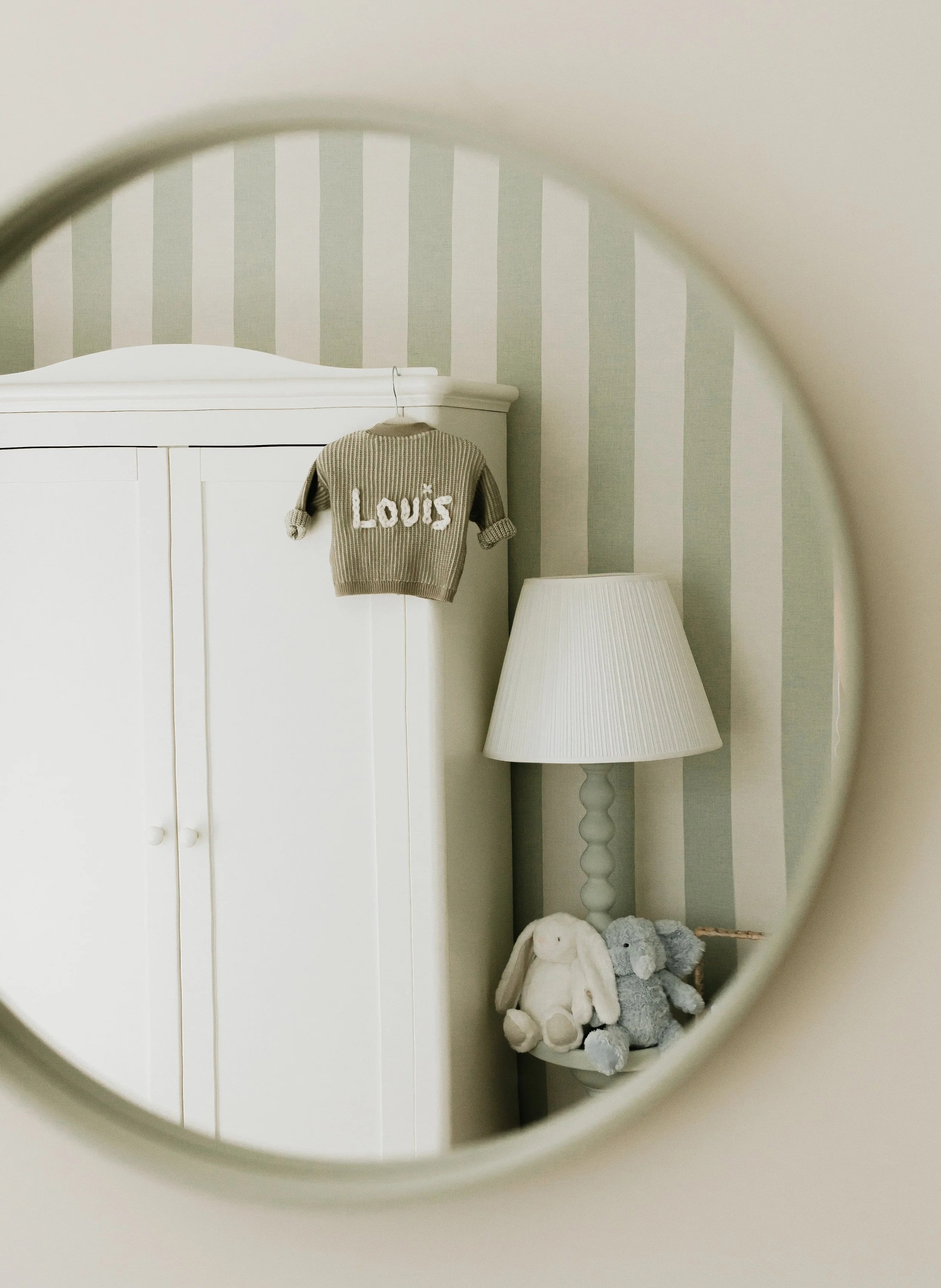 A white wardrobe with a small knit sweater hanging on the top, embroidered with the name 'Louis'. Next to it is a white lamp with a lampshade, and on the table are two plush stuffed animals, a white bunny and an elephant, all reflected in a mirror.