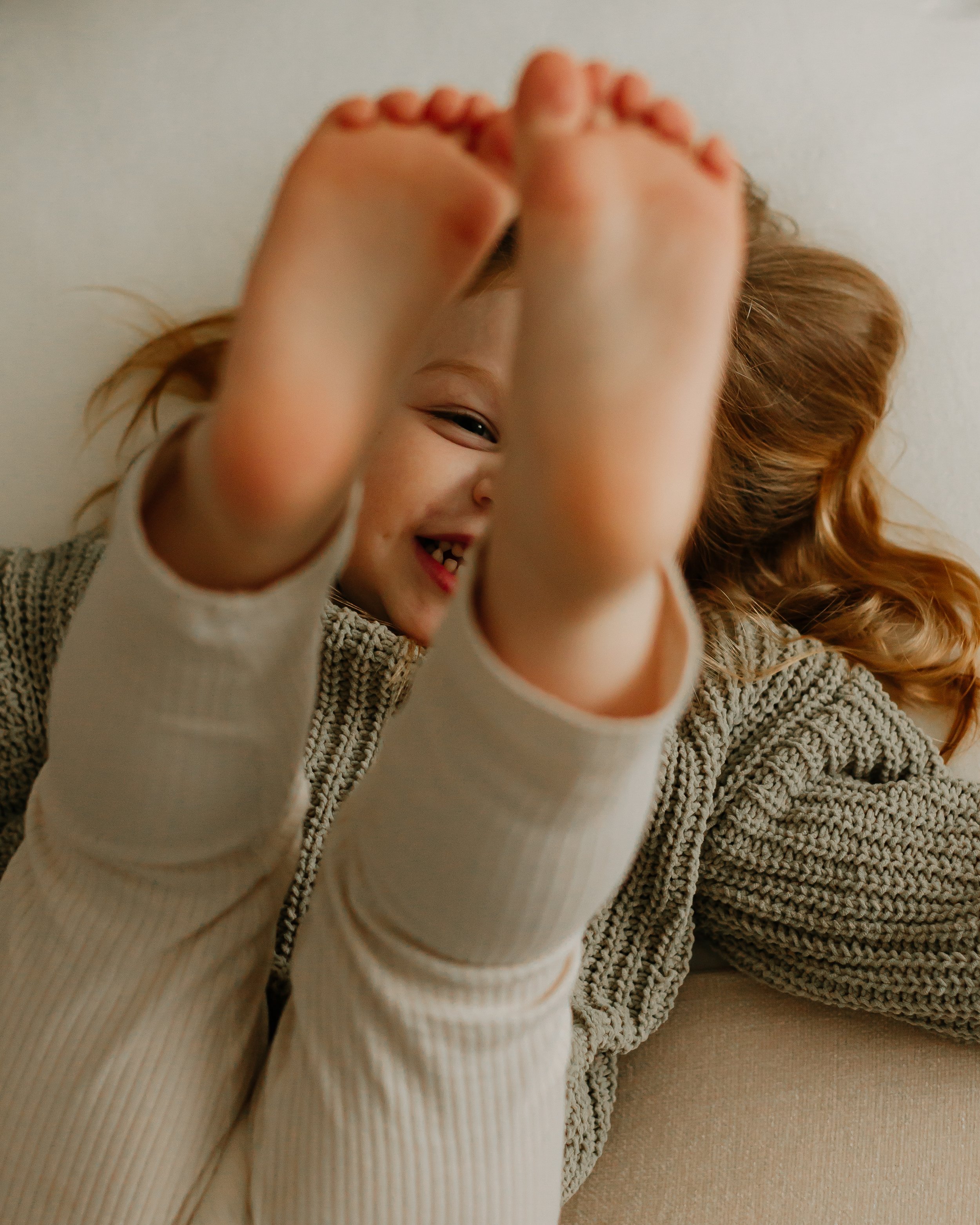 A young girl with brown hair smiling and lying on a couch, covering her face with her feet.