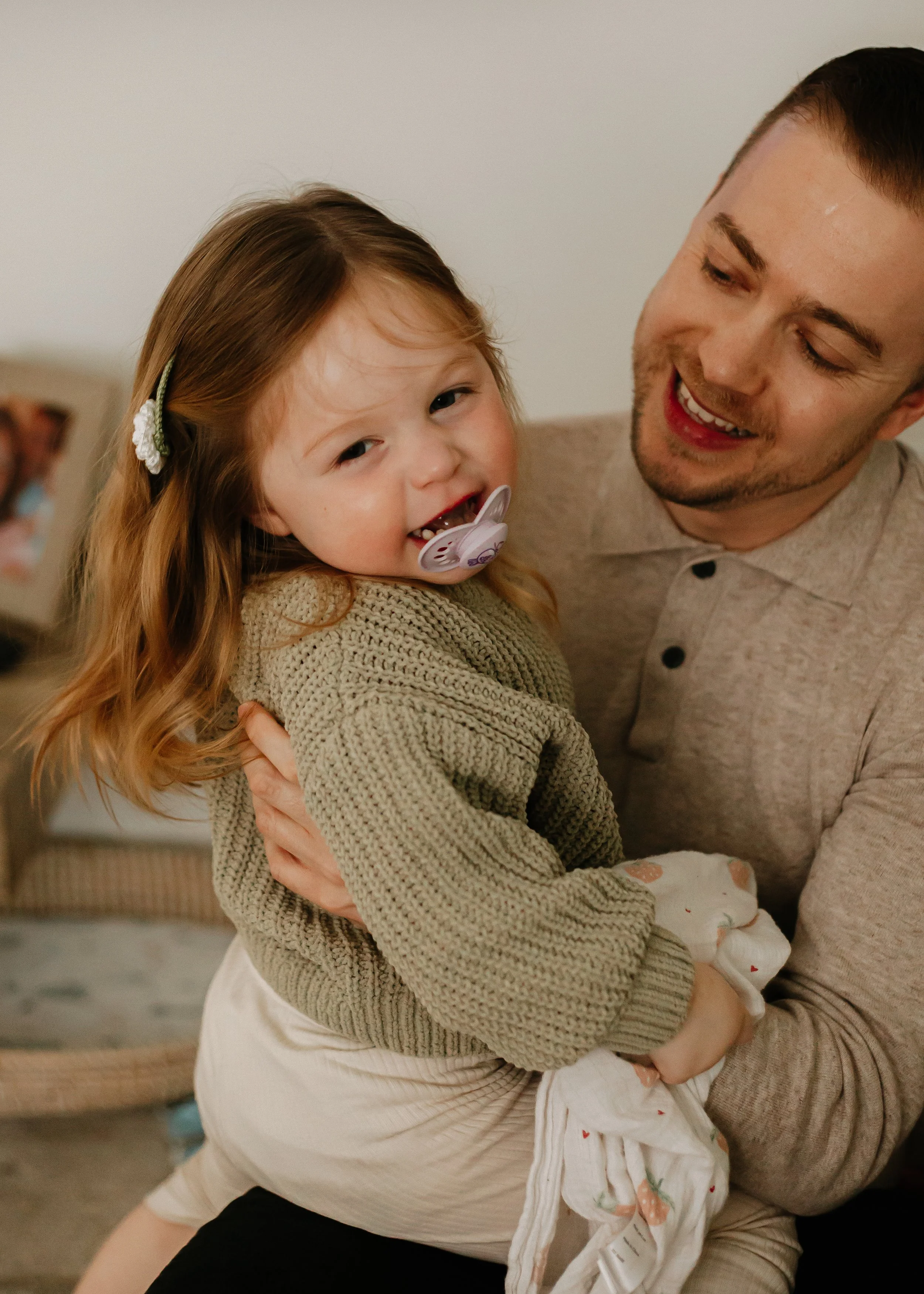 A young girl with brown and blonde hair and a pacifier in her mouth being held by a smiling man in a beige shirt.