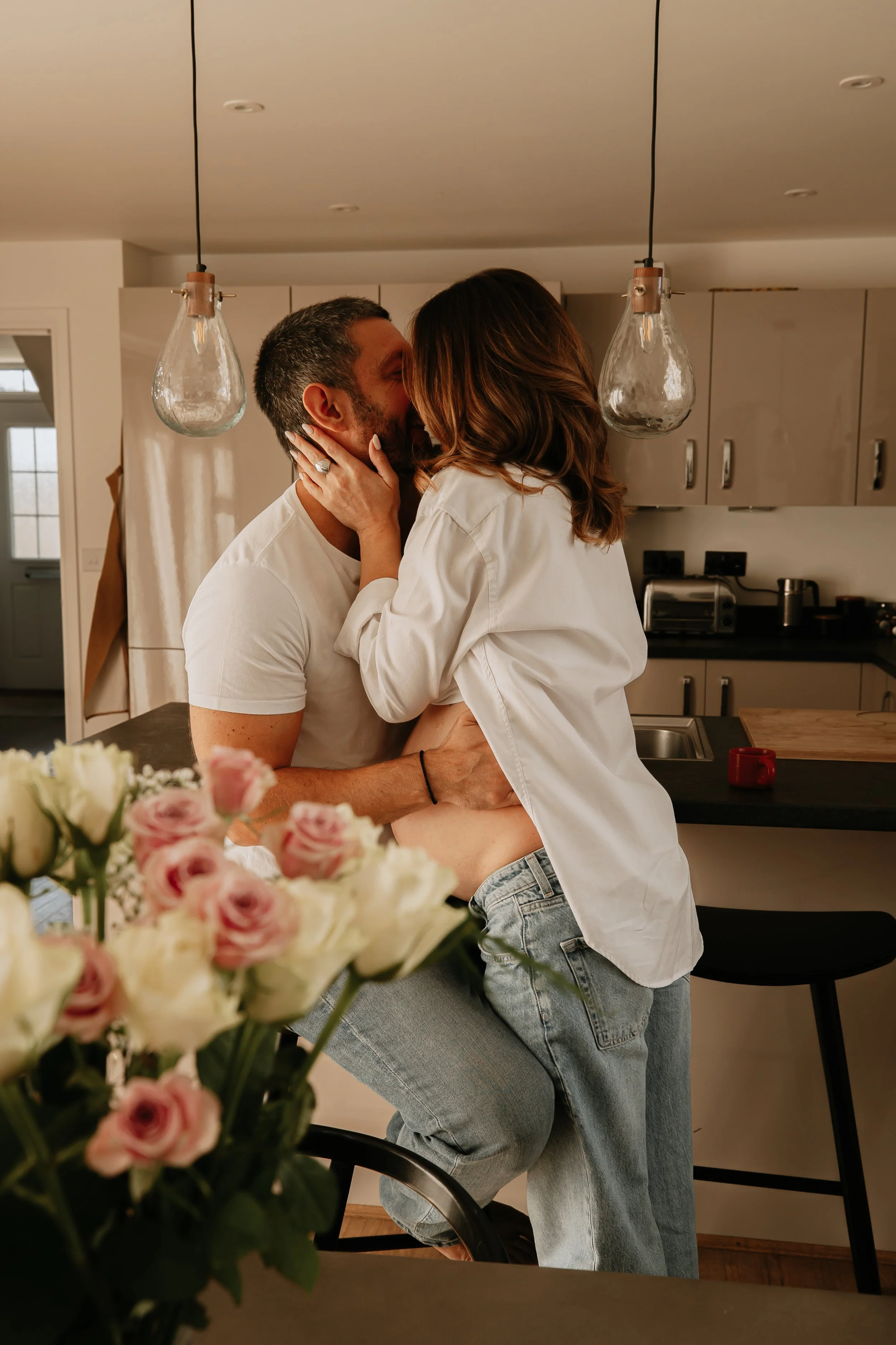 A man and pregnant woman sharing a kiss in a kitchen, with pink and white flowers in the foreground. 
