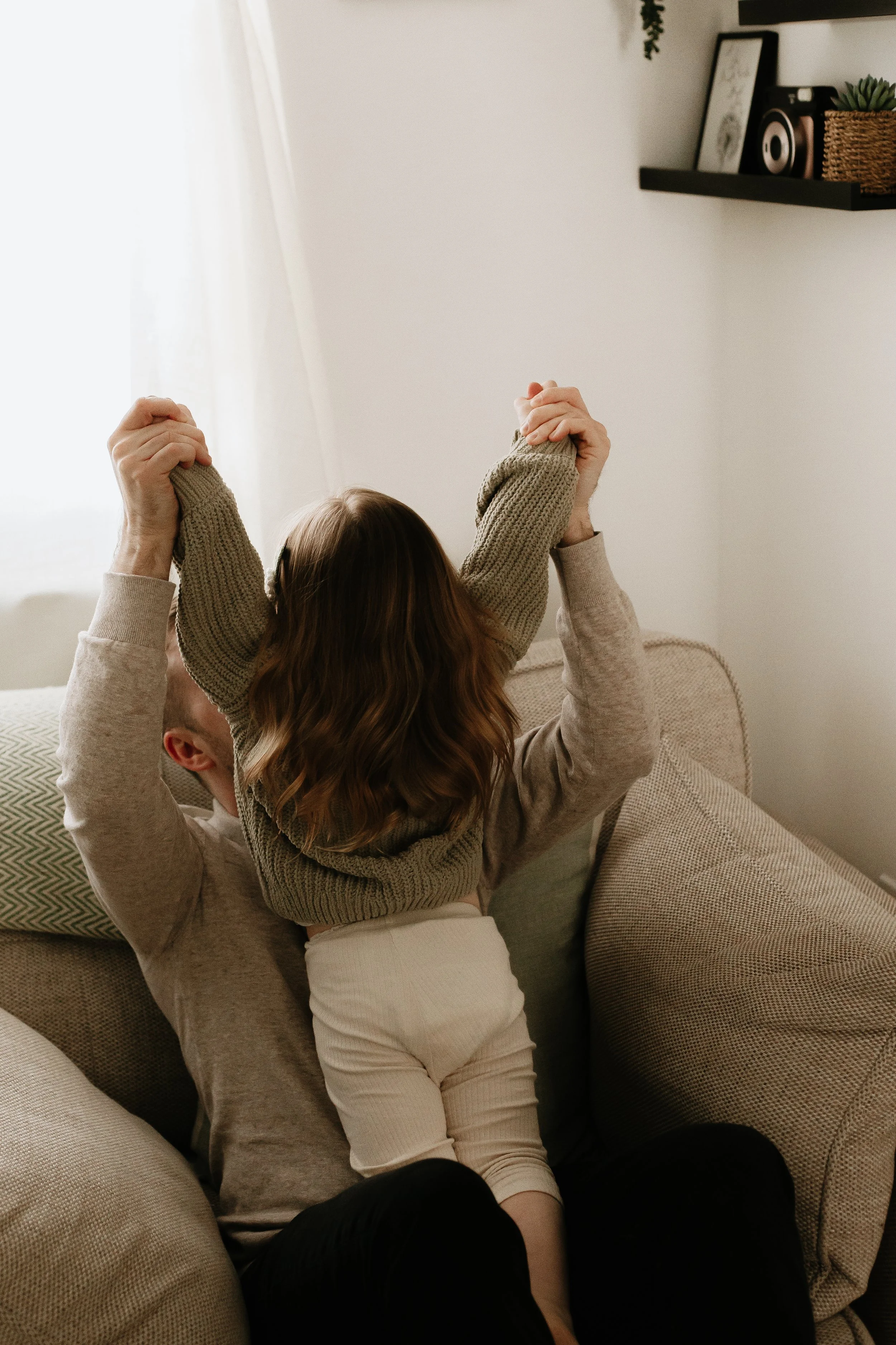 A man and a young girl stretch and play together on a beige couch in a living room with natural light and minimalist decor.