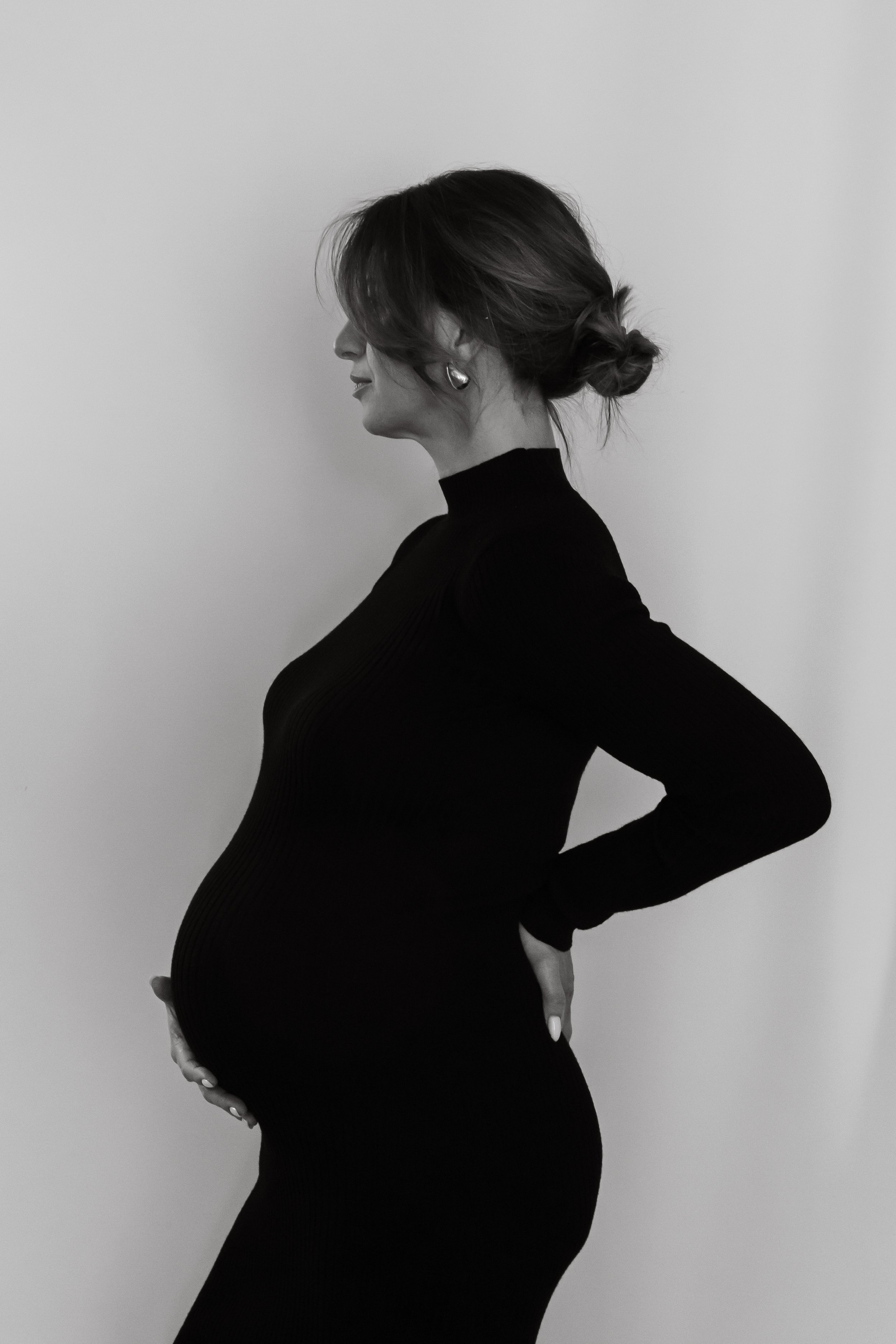 Black and white profile of a pregnant woman in a black turtleneck dress, with her hand resting on her back and her other hand on her belly.