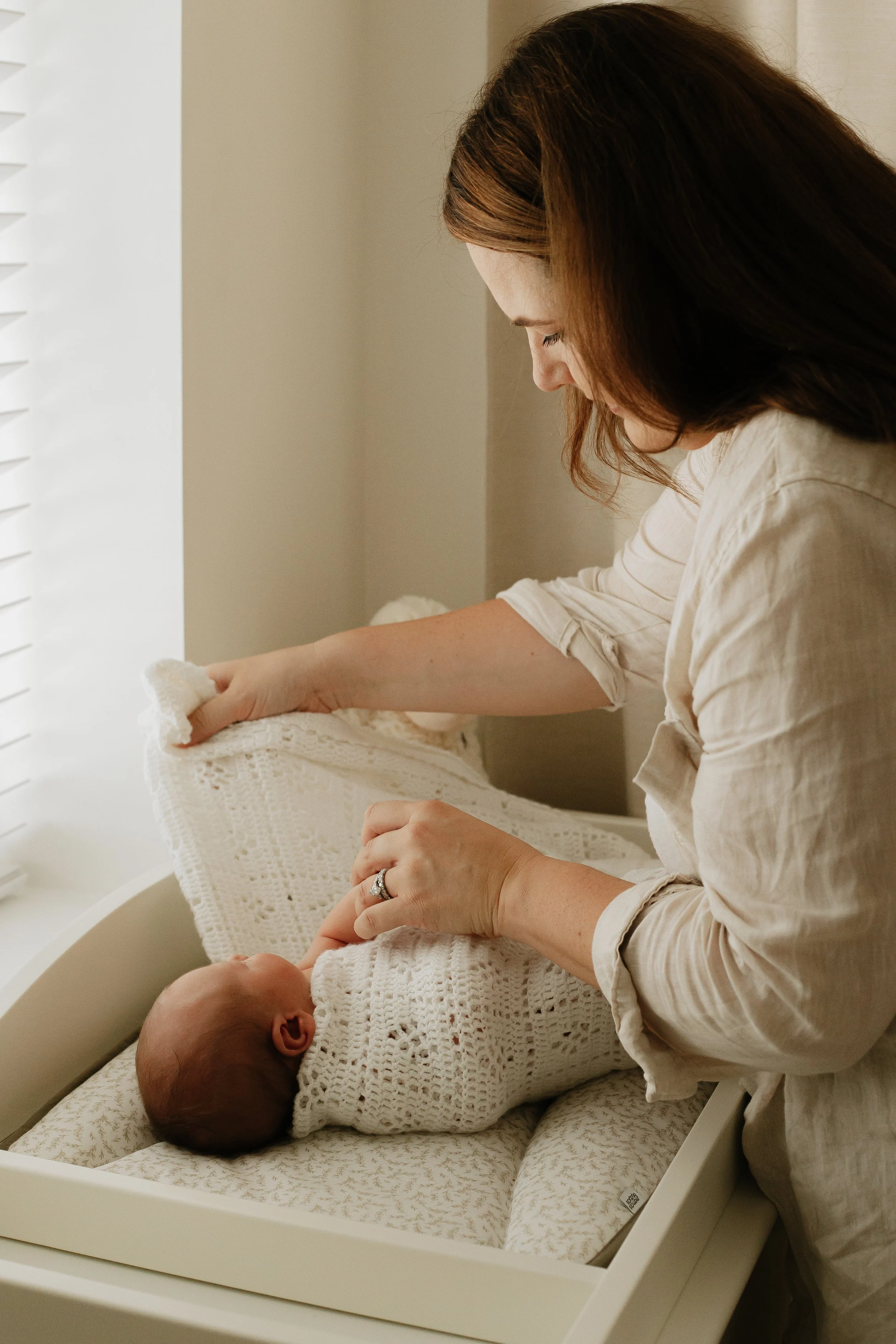 A woman gently lifting a white knitted blanket from a bassinet containing a newborn baby. The woman is wearing a beige shirt and has brown hair. The setting appears to be a softly lit room.