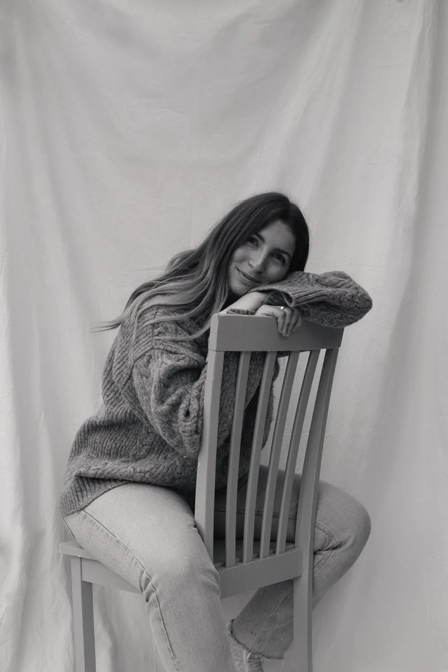 A young woman with long hair wearing a sweater and jeans, sitting sideways on a chair, resting her chin on her arm, smiling softly, against a light-colored background.