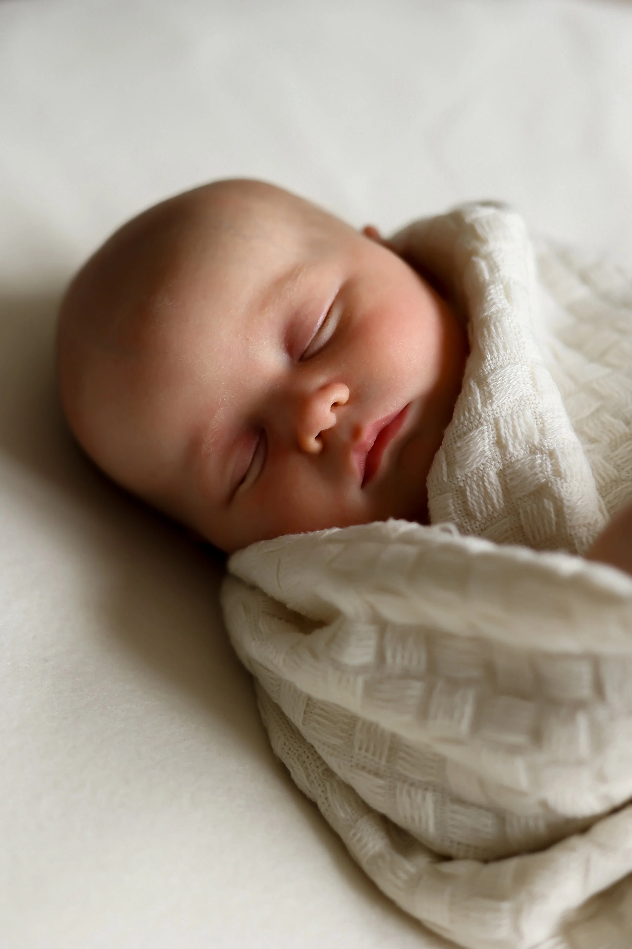 Close-up of a sleeping baby, peacefully resting on a white surface, wrapped in a cream-coloured textured blanket.