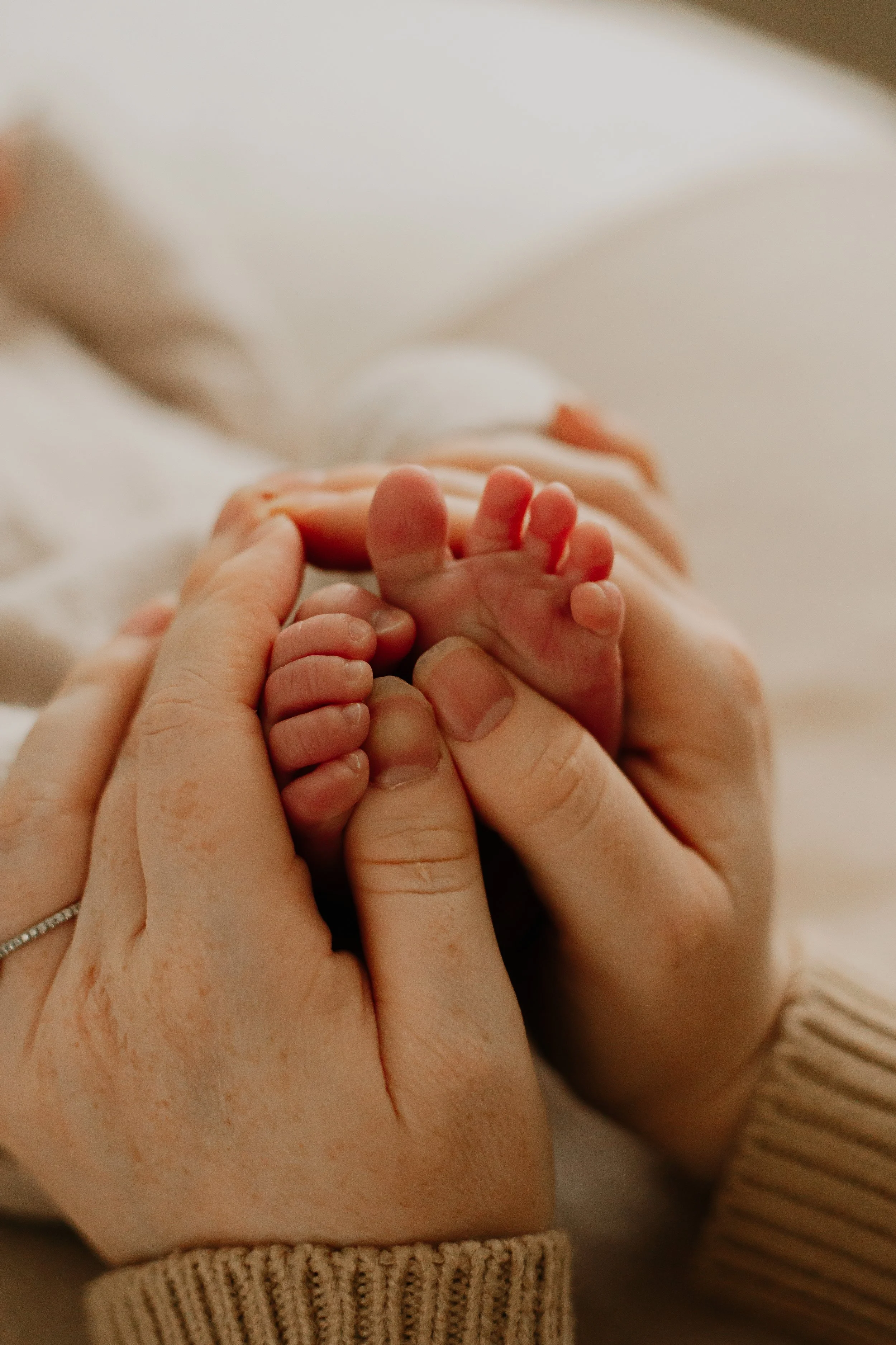 Adult's hands gently wrapped around baby's feet. The baby is very small with tiny toes, and the adult is wearing a beige sweater.