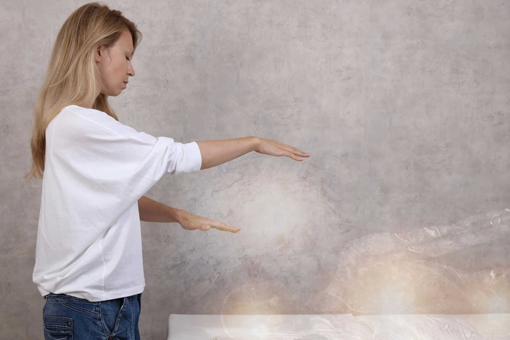 A woman in a white shirt and jeans is gesturing with her hands, with a neutral expression, in front of a textured wall.