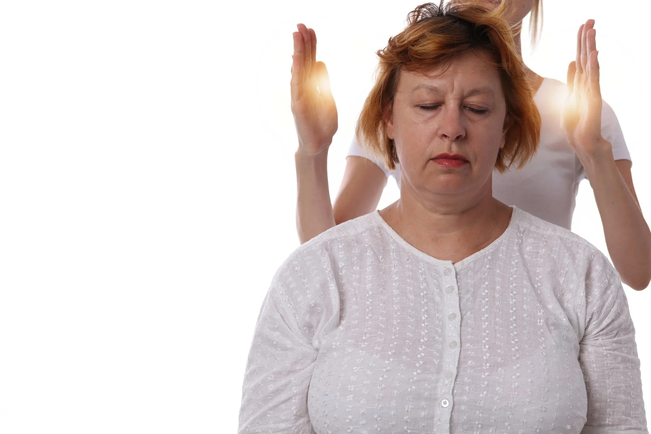 A woman with red hair in a white top appears to be meditating with her eyes closed, while another person stands behind her with their hands raised near her head, suggesting a massage or energy healing session.