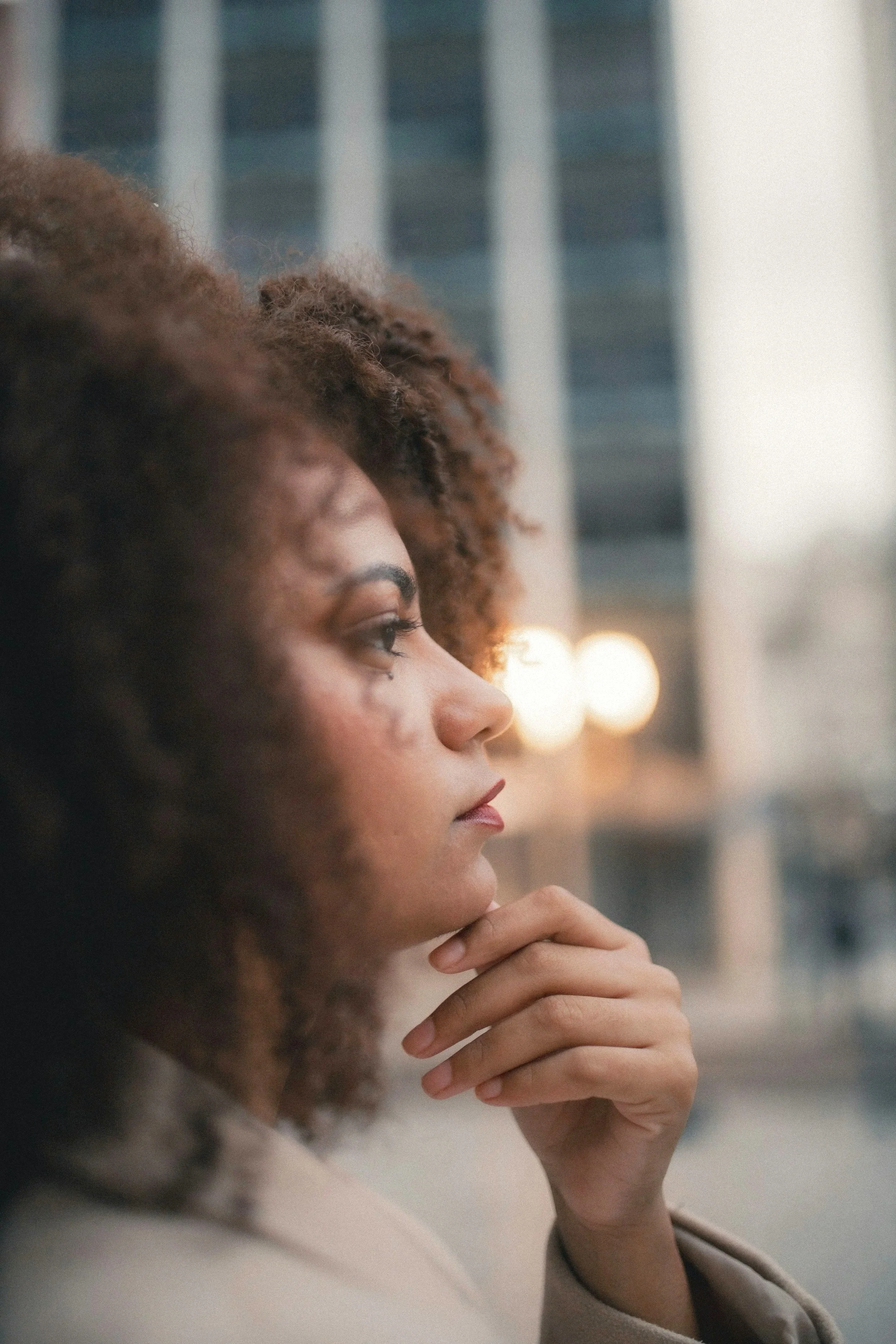 Close-up of a young woman's face in profile, with her hand resting on her chin, in front of a blurred cityscape with tall buildings and lights.