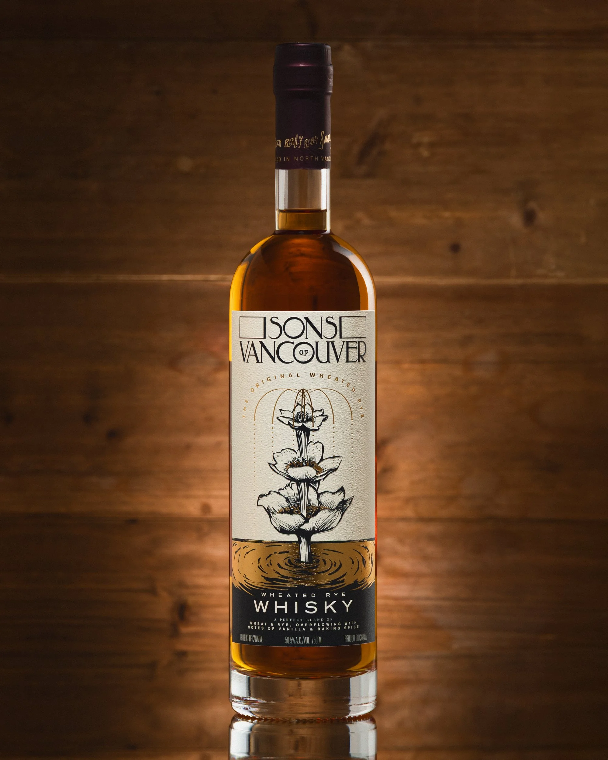 Bottle of Sons of Vancouver wheated rye whisky with a beige label and black and gold lettering, featuring an illustration of a layered flower with water ripples, set against a wooden background.