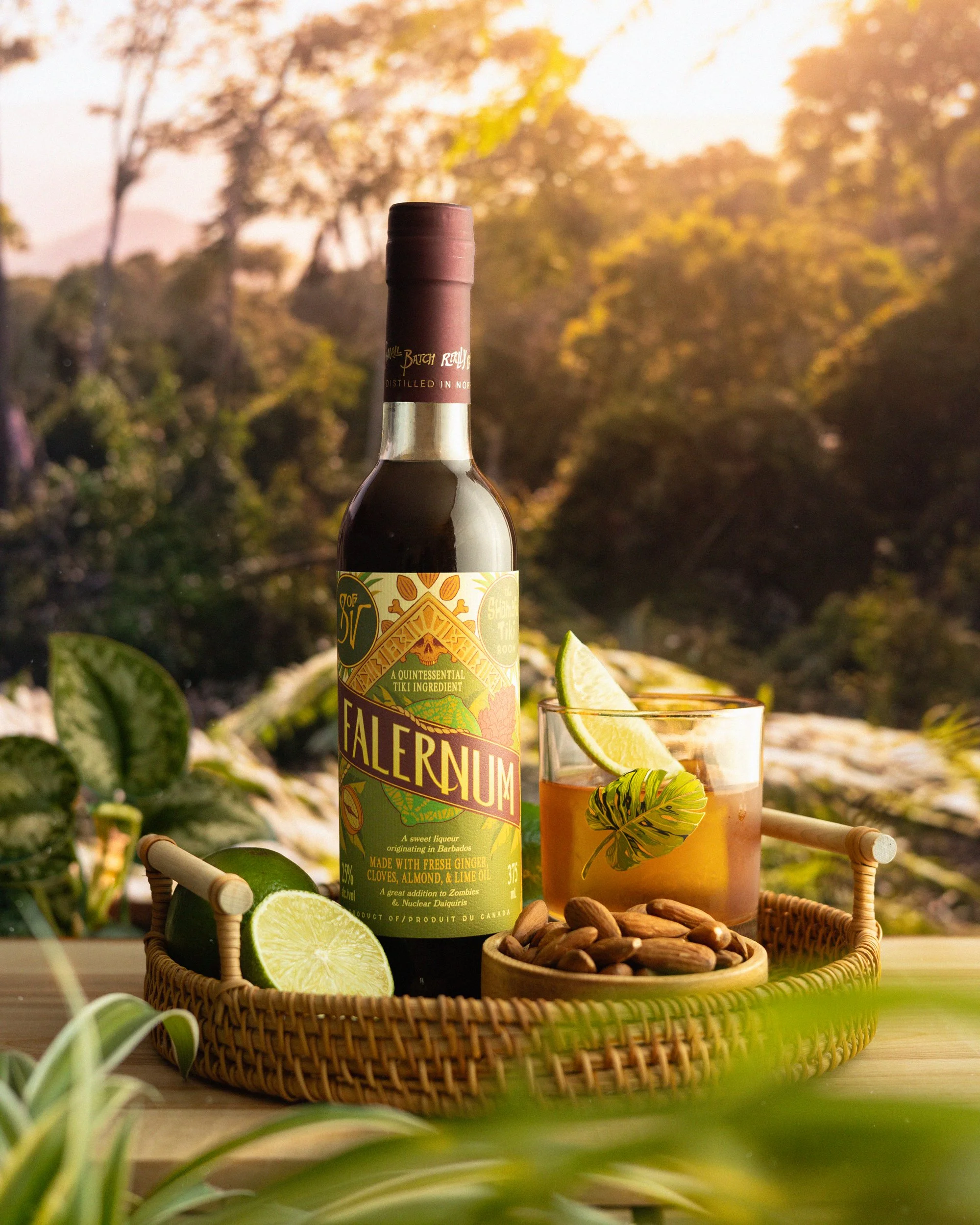 A bottle of Falernum liqueur with a glass of cocktail garnished with a lime wedge and a mint leaf, a bowl of almonds, and slices of lime on a woven tray outdoors with a background of trees and mountains at sunset.