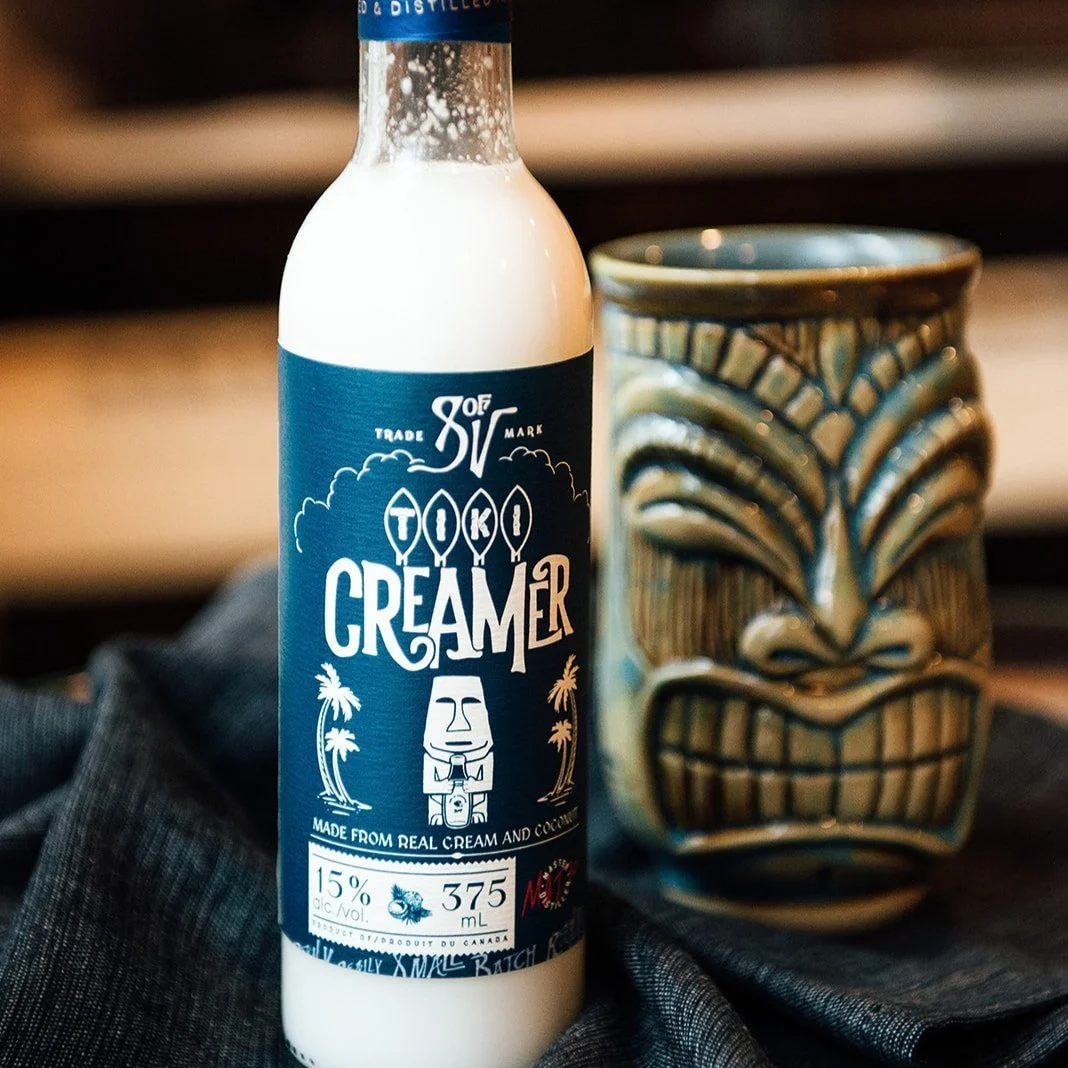 A bottle of tiki-themed cream liqueur labeled 'TIKI CREAMER' with a dark blue label featuring white graphics of palm trees and a tiki face, placed next to a decorative tiki mask on a black fabric surface.