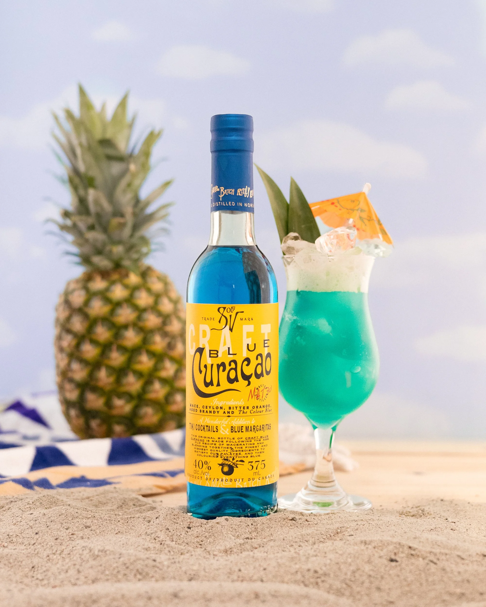 A bottle of Craft Curacao blue alcohol, a pineapple, and a blue cocktail with a pineapple leaf and small orange umbrella on a sandy beach.