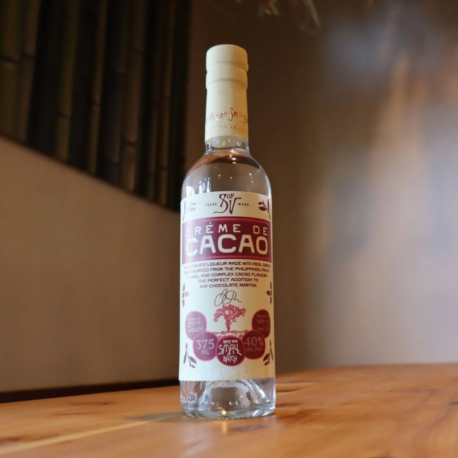 A clear bottle of cacao-flavored liqueur with a white cap, labeled 'Crème de Cacao,' sitting on a wooden surface against a blurred background.
