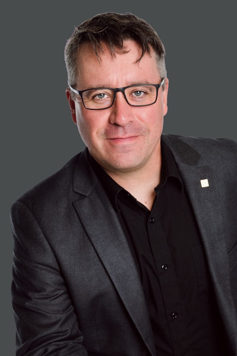 Homme d'affaires portant des lunettes, en costume gris et chemise noire, souriant, fond gris.