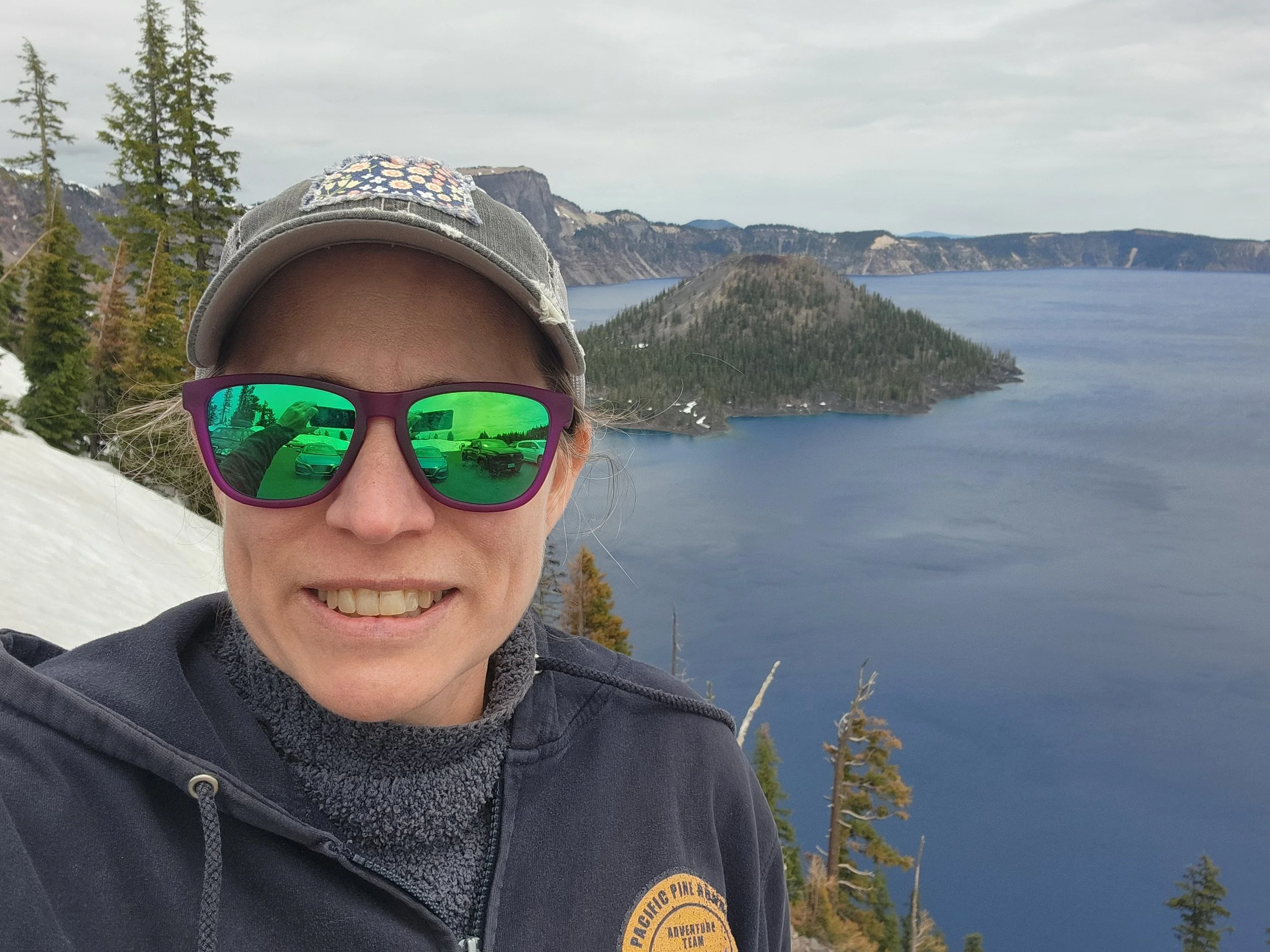 A smiling woman wearing sunglasses and a cap takes a selfie at a scenic lakeside with forested hills and a volcanic crater in the background.