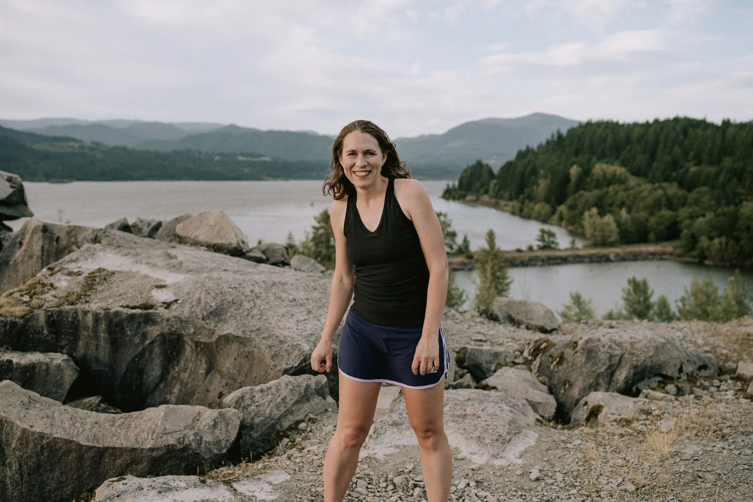A woman in a black tank top and navy shorts stands on rocky terrain near a large body of water with mountains and trees in the background.