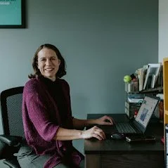 Woman smiling while sitting at a desk with a laptop, in an office setting.