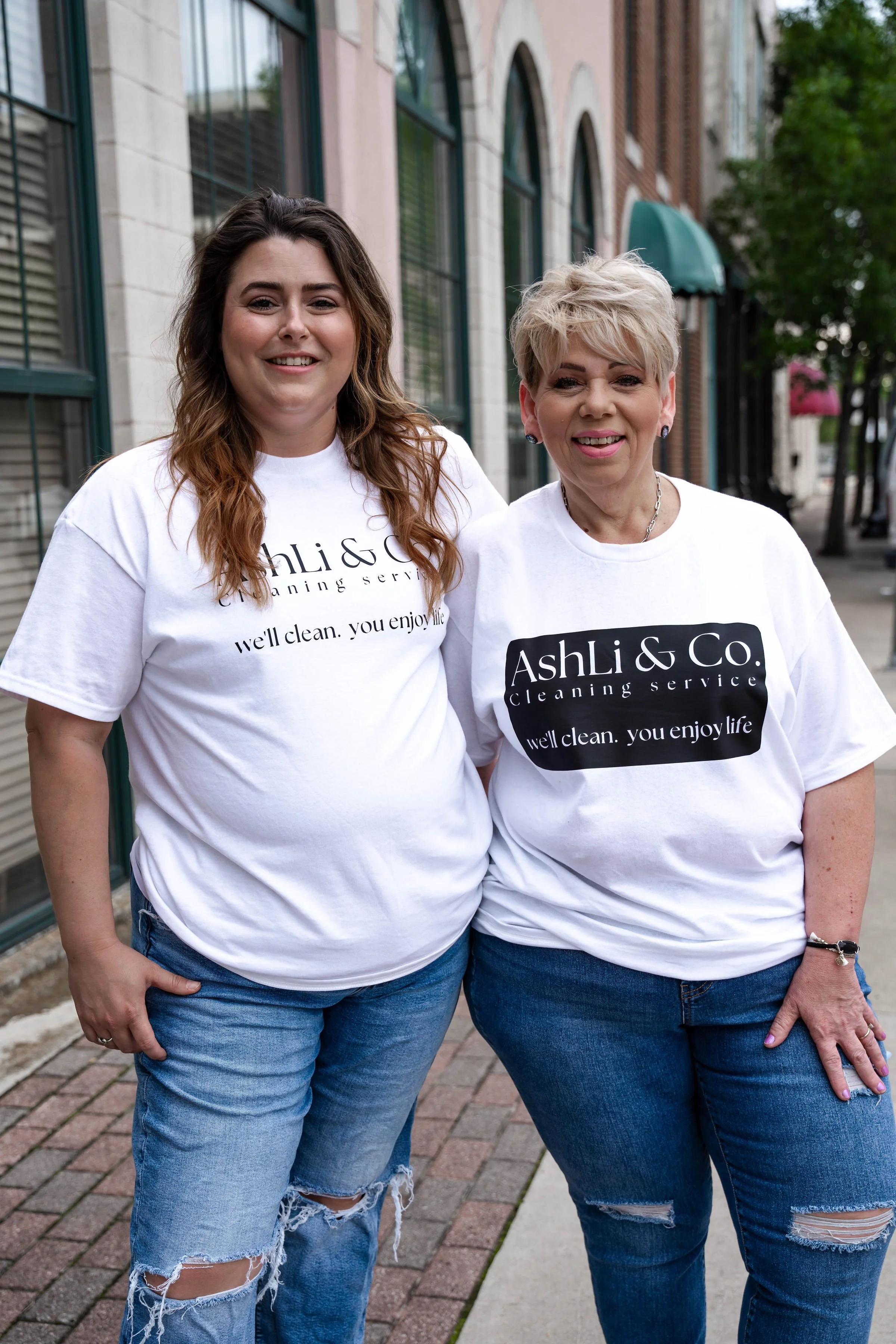 Two women standing side by side outdoors, both wearing white T-shirts with the text 'Ashli & Co. cleaning service' and 'well clean. you enjoy life.' They are smiling, with the woman on the left having long, wavy brown hair, and the woman on the right having short, blonde hair. They are dressed in casual jeans, with a brick sidewalk and buildings with large windows and arched doorways in the background.
