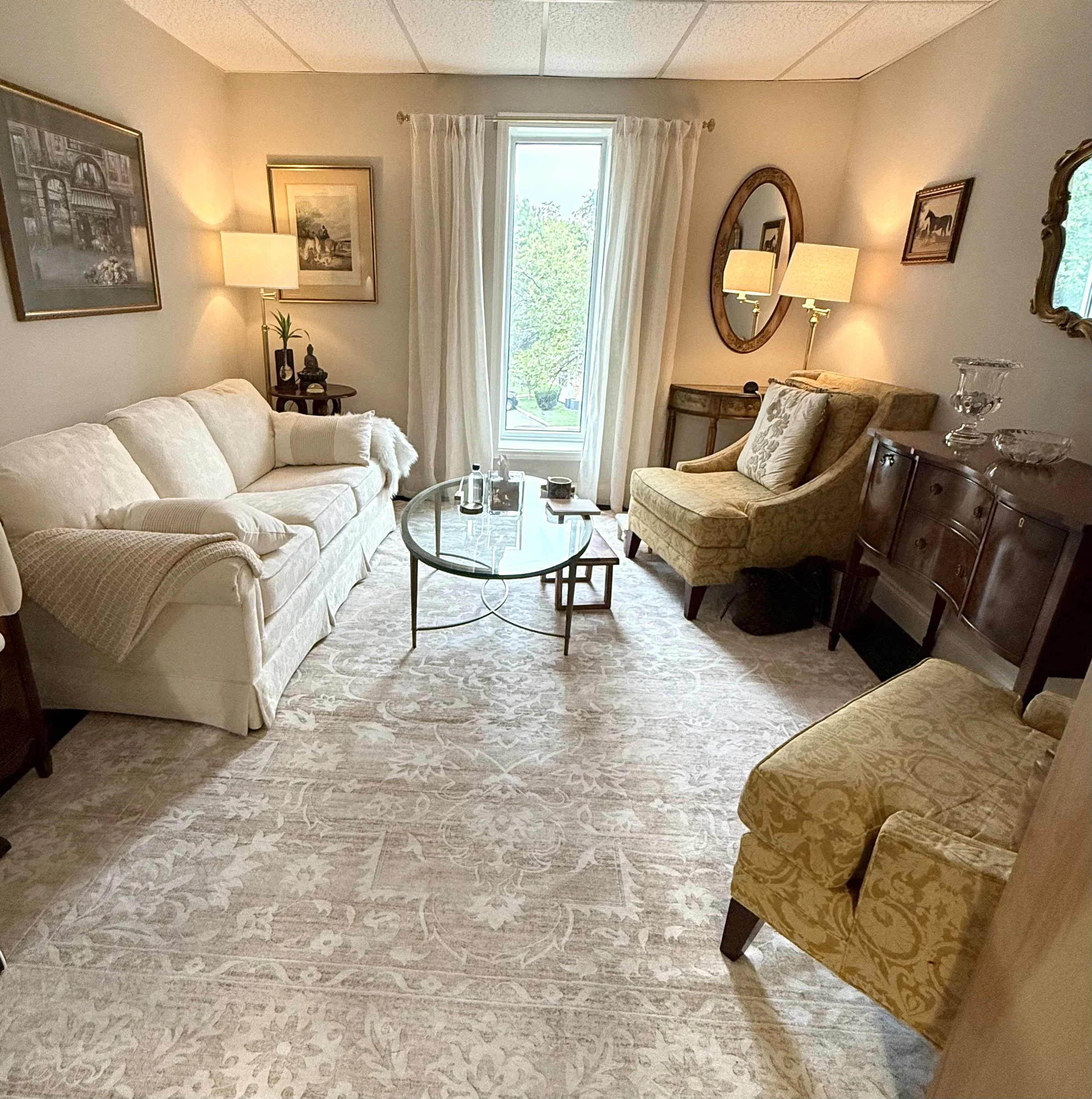 A cozy living room with a cream-colored sofa, an armchair with a pillow, a glass coffee table, framed artwork on the walls, a window with white curtains, and various lamps providing warm lighting.