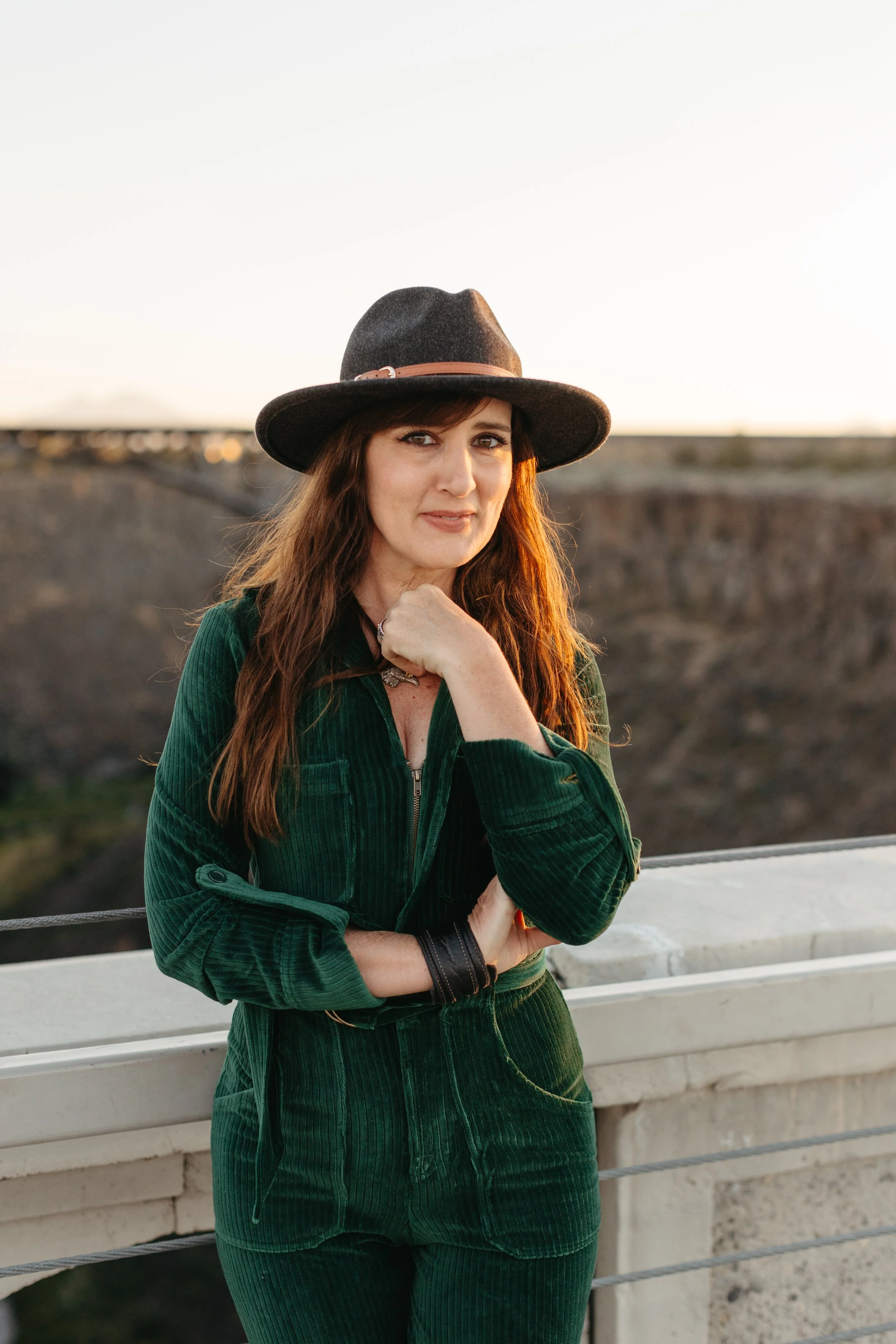A woman with long reddish-brown hair, wearing a black wide-brimmed hat with a pink band, a dark green corduroy jacket, and matching green pants, standing outdoors on a bridge during sunset, with a blurred landscape in the background.