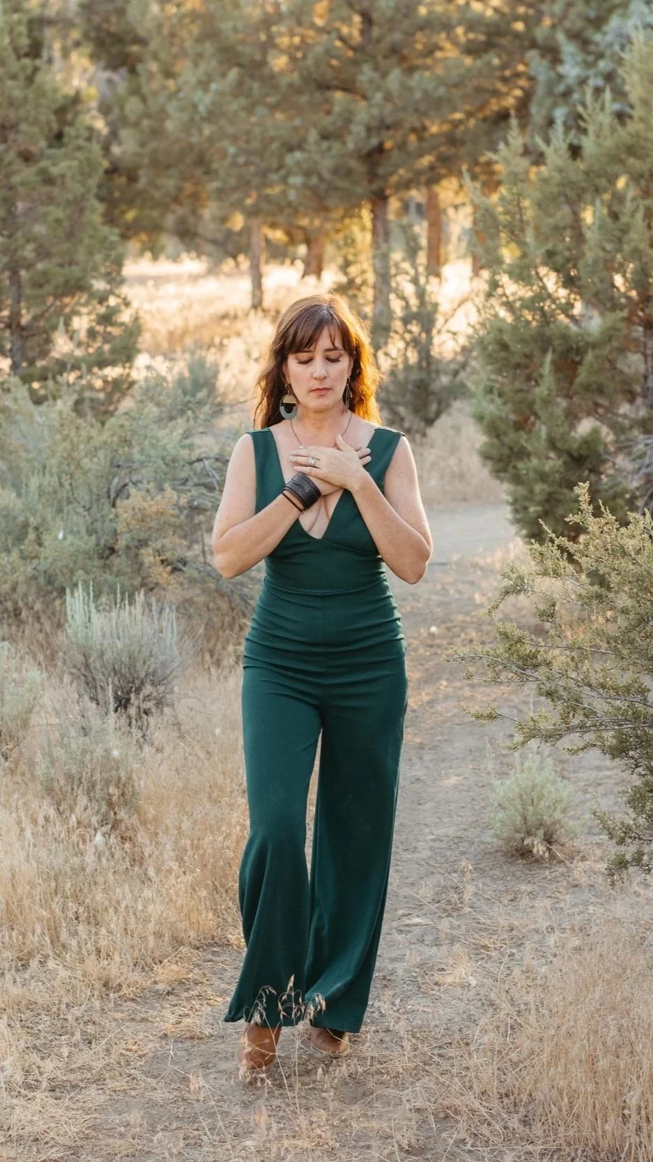 A woman in a dark green dress with a deep neckline is walking barefoot on a dirt trail in a dry, bushy landscape with trees. She is placing her hands over her chest, eyes closed, appearing to be in a meditative or prayerful moment, during sunset or golden hour.