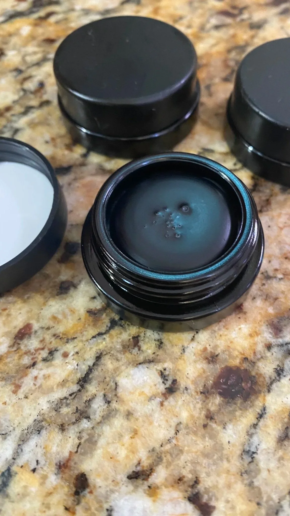 Open black jar with natural mascara on a granite countertop, surrounded by three closed similar jars.