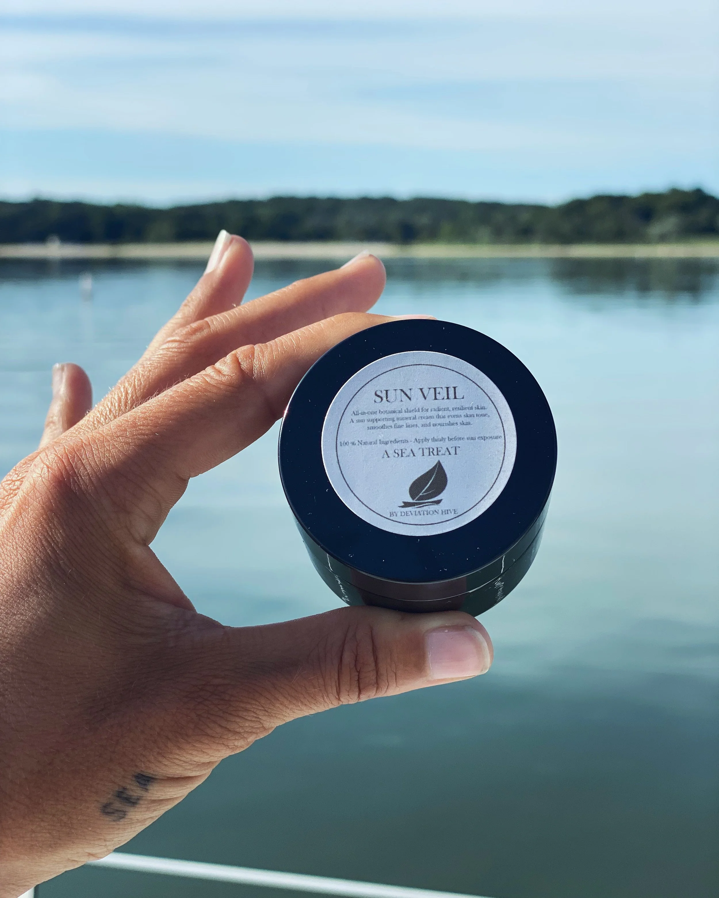 A person's hand holding a round container of Sun Veil natural sunscreen against a blurred body of water and sky in the background.
