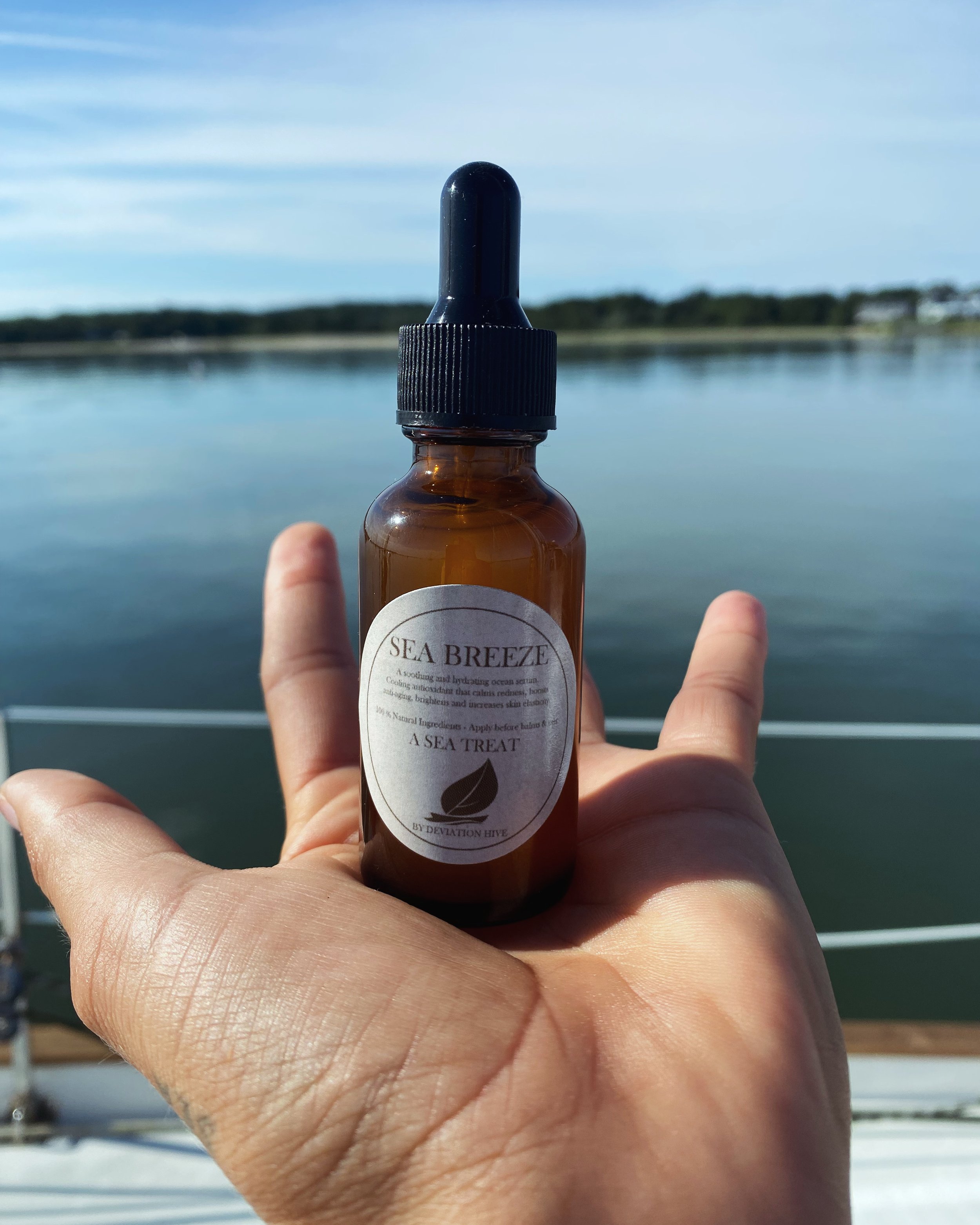 A person holding a small brown glass dropper bottle labeled 'Sea Breeze' with a dropper cap, against a backdrop of a body of water and a blue sky with clouds.