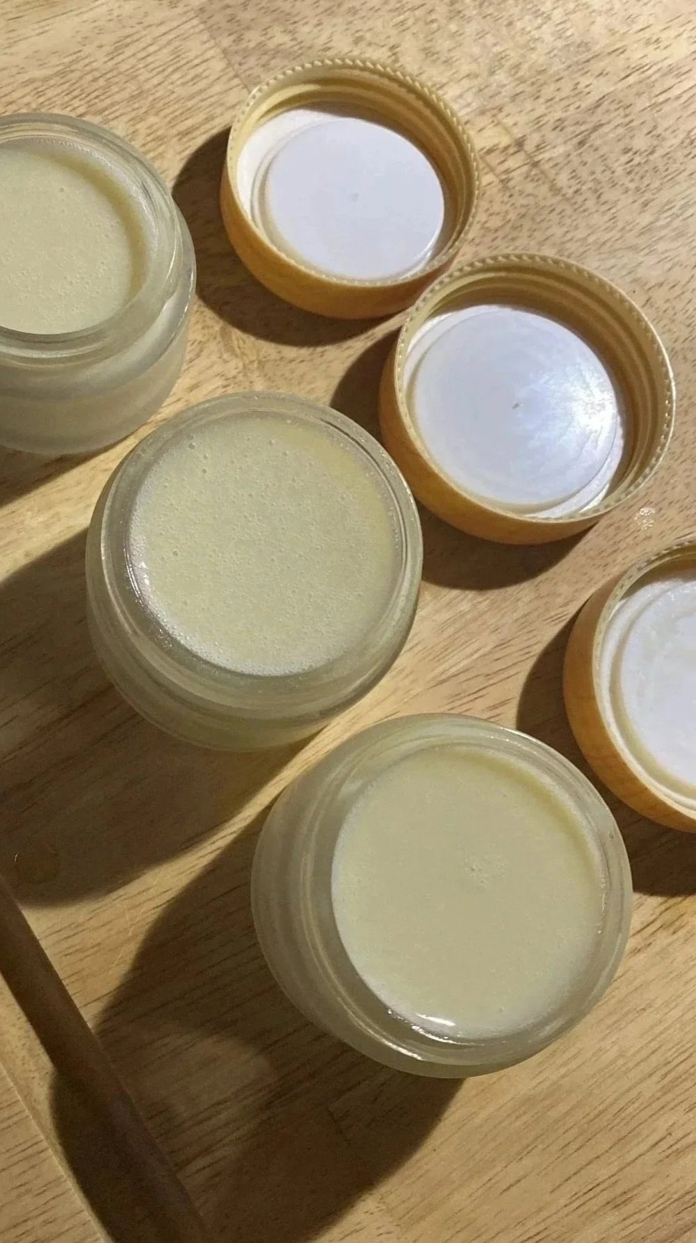 Multiple small glass jars filled with natural balms and deodorant cream, some with open lids, on a wooden surface.