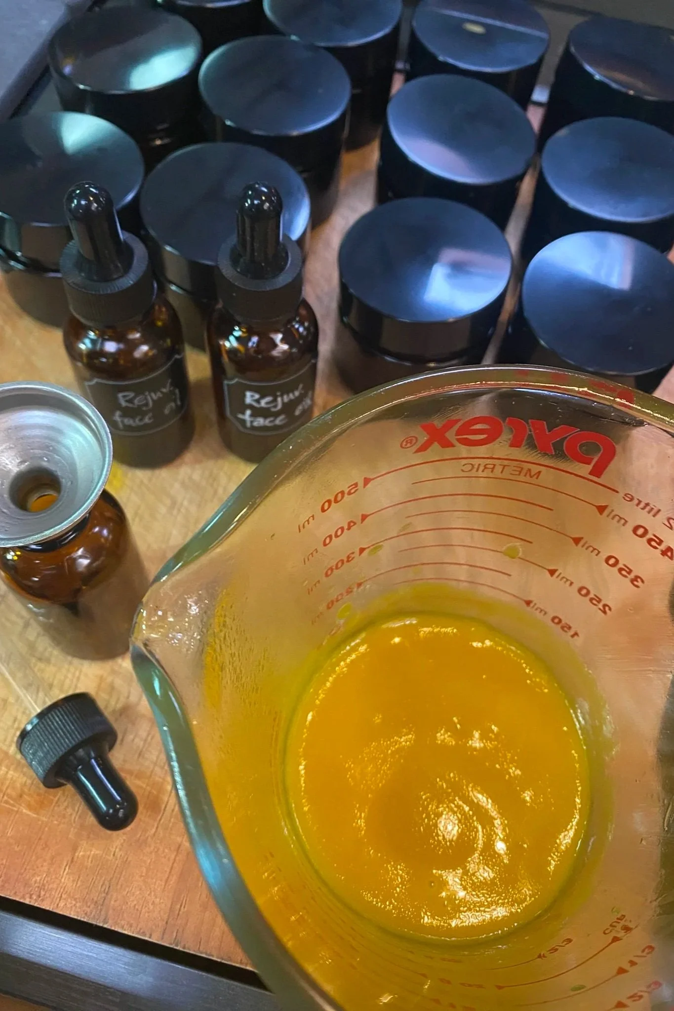 Various bottles and containers, including a measuring cup with a batch of sea glow, a sun support balm, a small dropper bottle labeled "Rejuvenate Face" and a small vial, are placed on a wooden surface, likely in a laboratory or skincare setting.